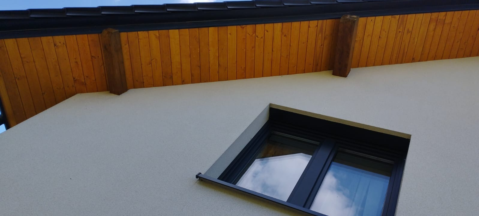 Fragment elewacji domu z oknem i drewnianą podbitką dachową z widocznymi belkami. Czarna rynna wzdłuż krawędzi dachu. Jasny tynk strukturalny.