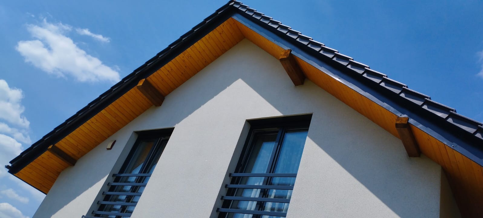 Nowoczesny dom z ciemną dachówką i drewnianą podbitką, widziany z dołu na tle błękitnego nieba. Wyraźne okna z czarnymi ramami i metalowymi barierkami.