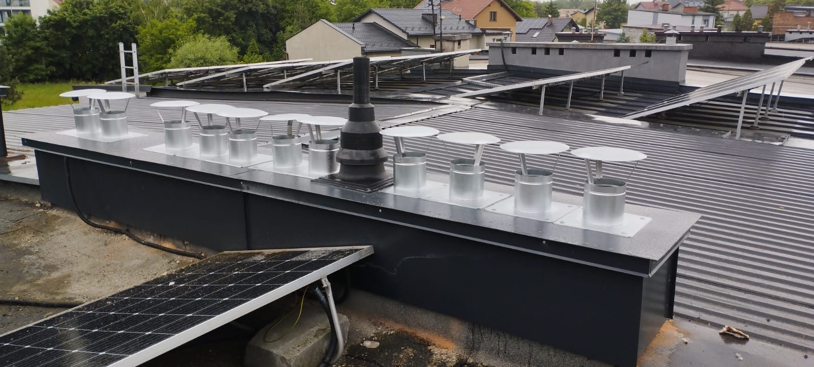 Płaski dach z blachy trapezowej, liczne kominki wentylacyjne z kołpakami i panele słoneczne. Widok z góry na szare tło budynków i zieleni.