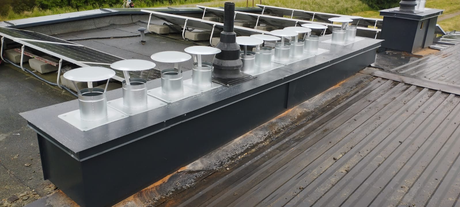 Szary dach z panelami słonecznymi i szeregiem metalowych kominków wentylacyjnych z daszkami, ustawionych na podwyższeniu. Widoczne zabrudzenia na powierzchni.