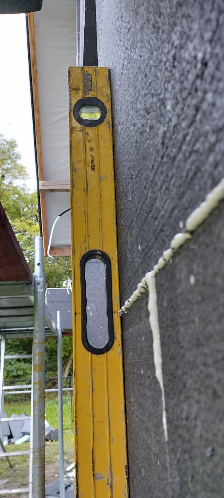 Poziomica przyłożona do szarej ściany z ociepleniem, widoczne fragmenty rusztowania i dachu. Prace dekarskie, ocieplanie budynku.