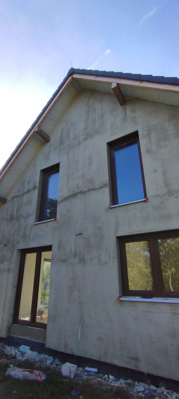 Surowy budynek jednorodzinny z ciemnymi oknami i dachem z dachówki ceramicznej. Elewacja w trakcie prac, widoczne instalacje elektryczne. Ujęcie z dołu, niebo w tle.