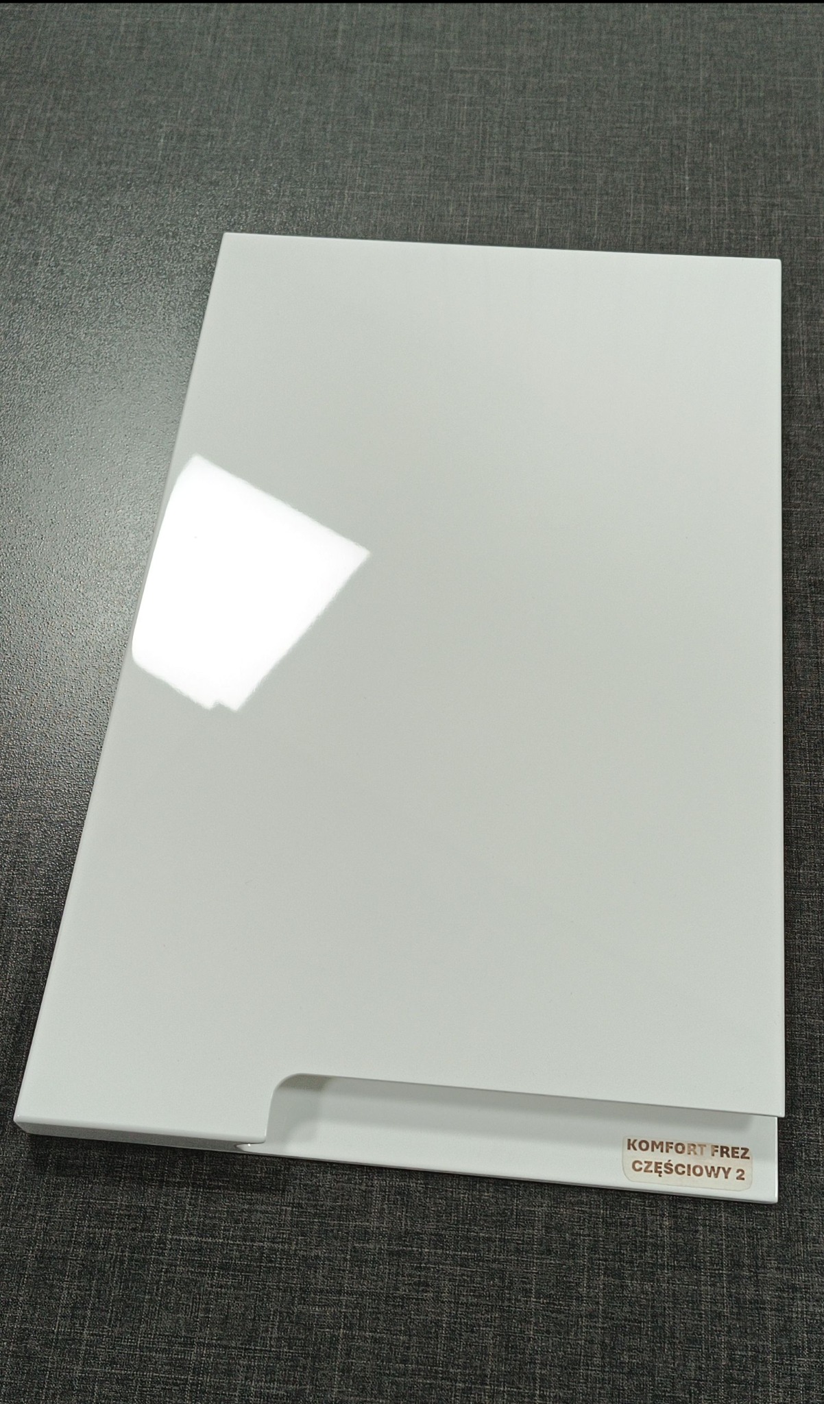 Biały, lakierowany front meblowy z frezowanym uchwytem, leżący na ciemnoszarym blacie z widoczną naklejką 'Komfort Frez Częściowy 2'.