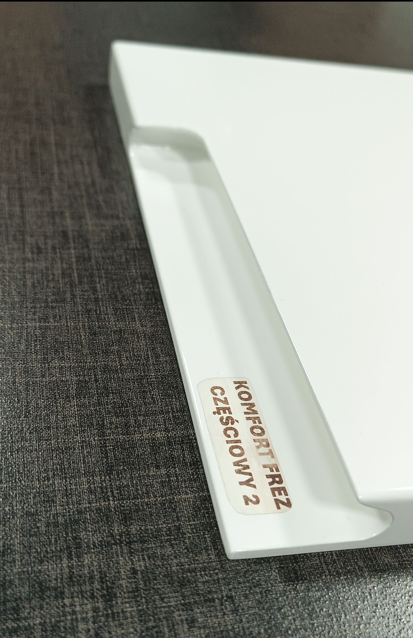 Biały element wykończeniowy z naklejoną etykietą 'KOMFORT FREZ CZĘŚCIOWY 2' leżący na szarej powierzchni.