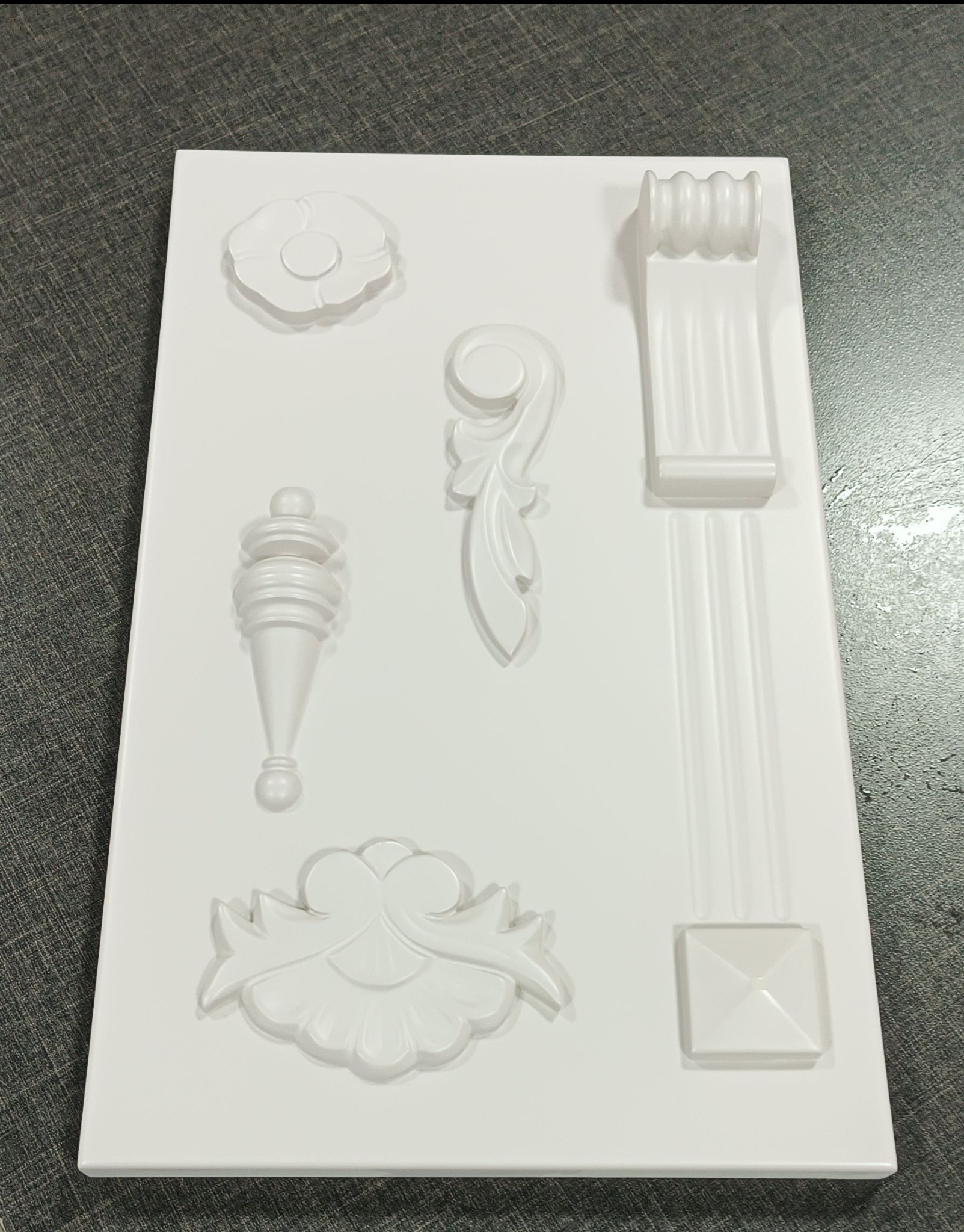 Biała, plastikowa forma z odlewami dekoracyjnych elementów architektonicznych, w tym rozety, kapitele i ornamenty roślinne, na szarym tle.