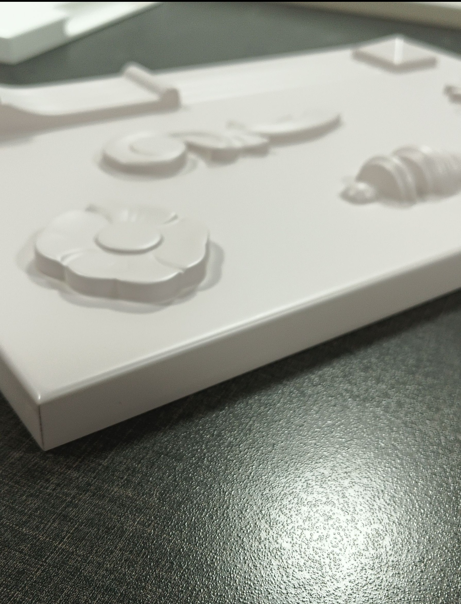 Biała płyta z wytłoczonymi wzorami kwiatów, spirali i geometrycznych kształtów, leżąca na ciemnej, teksturowanej powierzchni.