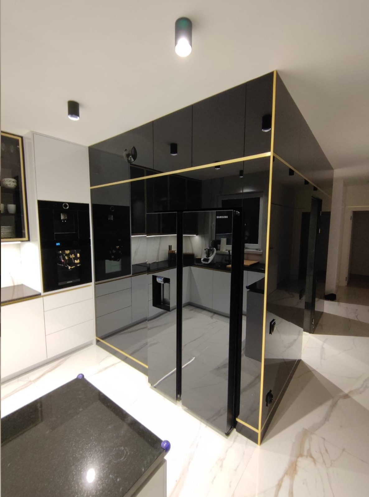 Nowoczesna kuchnia z czarnymi, lakierowanymi frontami szafek, złotymi akcentami i marmurową podłogą, oświetlona punktowymi lampami sufitowymi.