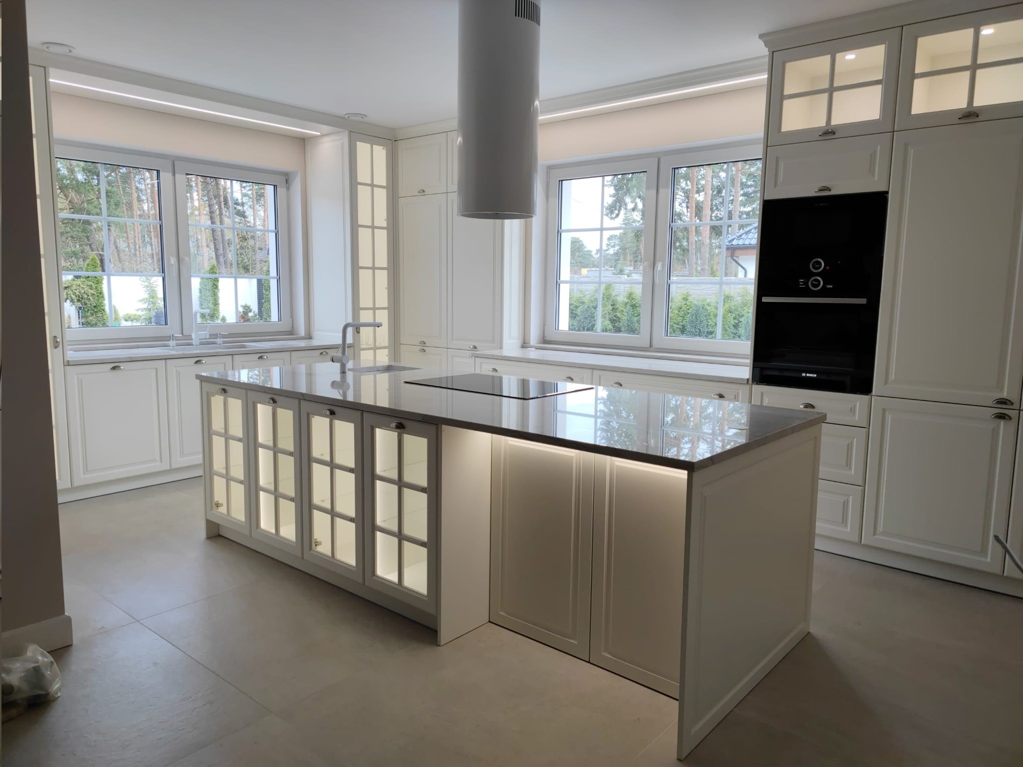 Jasna, przestronna kuchnia z wyspą, białe meble z frezowanymi frontami, szklane witryny w wyspie podświetlone od wewnątrz, okap cylindryczny, duże okna z widokiem na zieleń.