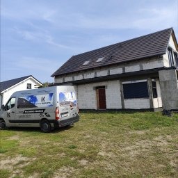 Dom w trakcie budowy z szarą dachówką, widoczny z poziomu trawnika, z samochodem dostawczym firmy budowlanej na pierwszym planie i błękitnym niebem w tle.