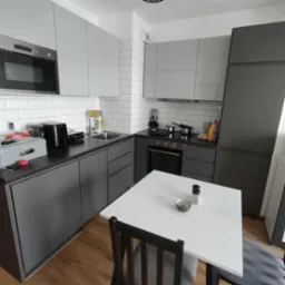 Nowoczesna, szaro-biała kuchnia z białymi płytkami na ścianie, czarnym blatem i drewnianą podłogą. Na stole miseczka i przyprawy.