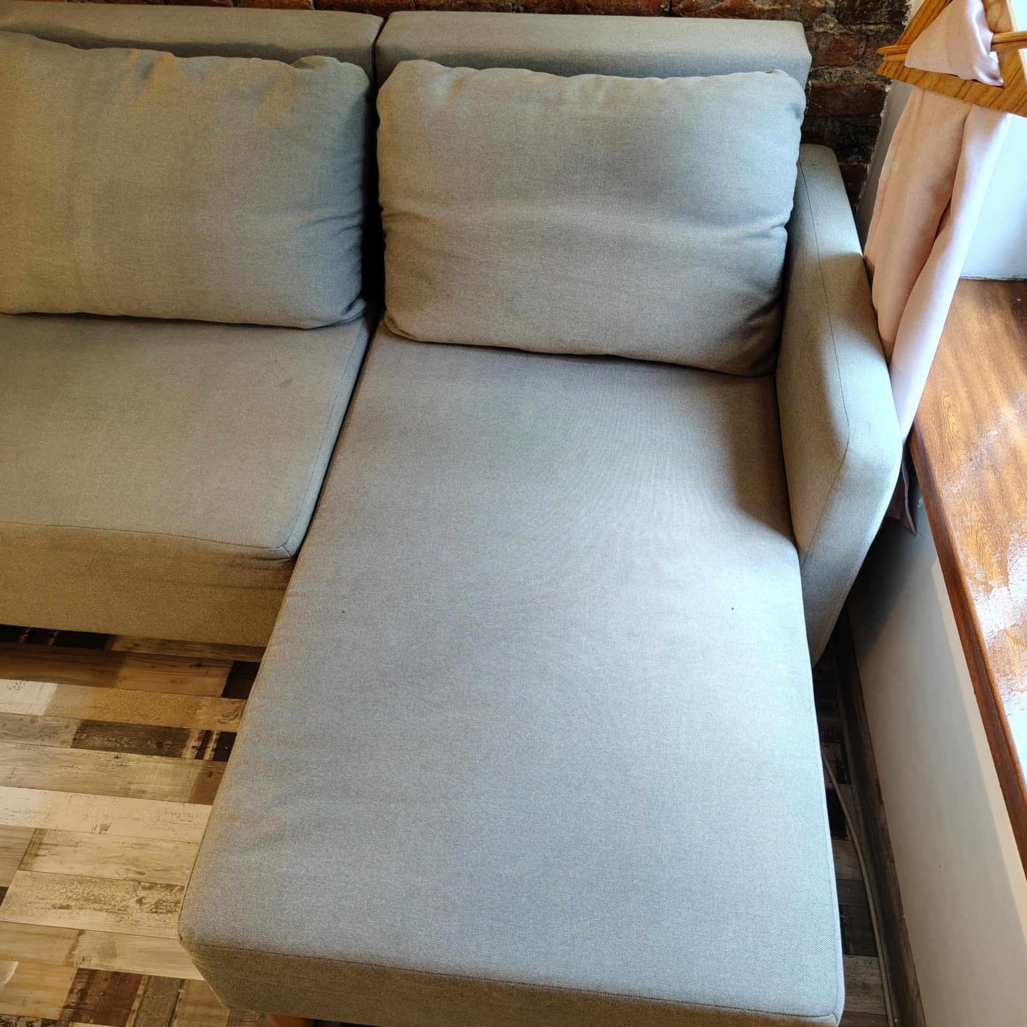 Jasnoszara kanapa z poduszkami, widoczna z góry, z drewnianą podłogą i fragmentem ceglanej ściany w tle. Na oparciu wieszak z jasną tkaniną.