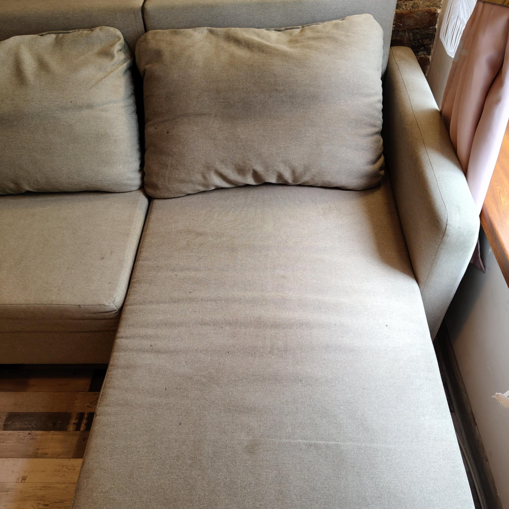 Beżowa kanapa z poduszkami, widoczne plamy na siedzisku. W tle drewniana ściana i fragment okna z zasłoną. Fotografia z bliska, ukazująca detale tapicerki.