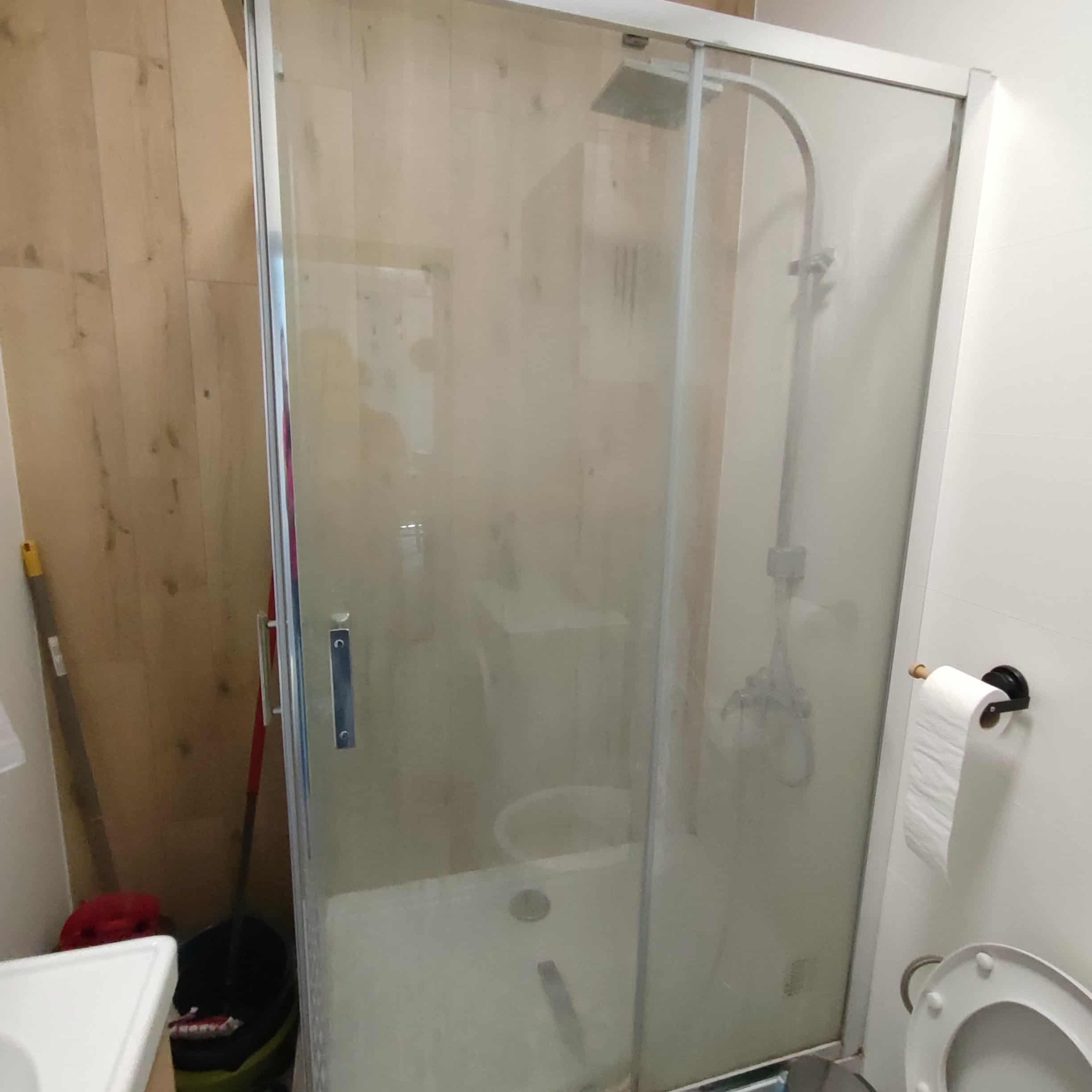 Kabina prysznicowa po czyszczeniu, widoczne zacieki na szybie. W tle drewniana ściana i akcesoria toaletowe. Ujęcie z bliska ukazuje detale.