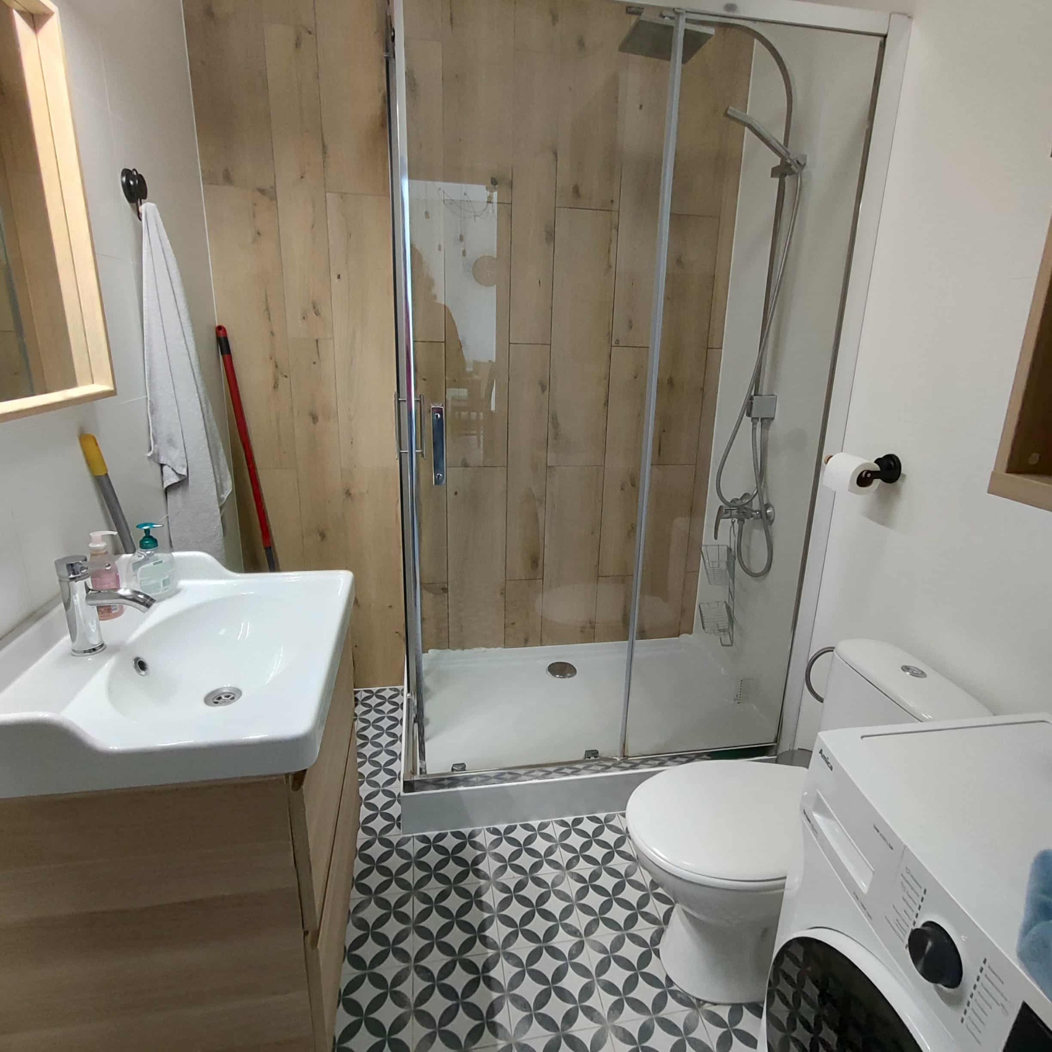 Umywalka z szafką, toaleta, kabina prysznicowa i pralka w małej łazience z drewnianymi panelami i wzorzystą podłogą. Widoczne akcesoria łazienkowe.