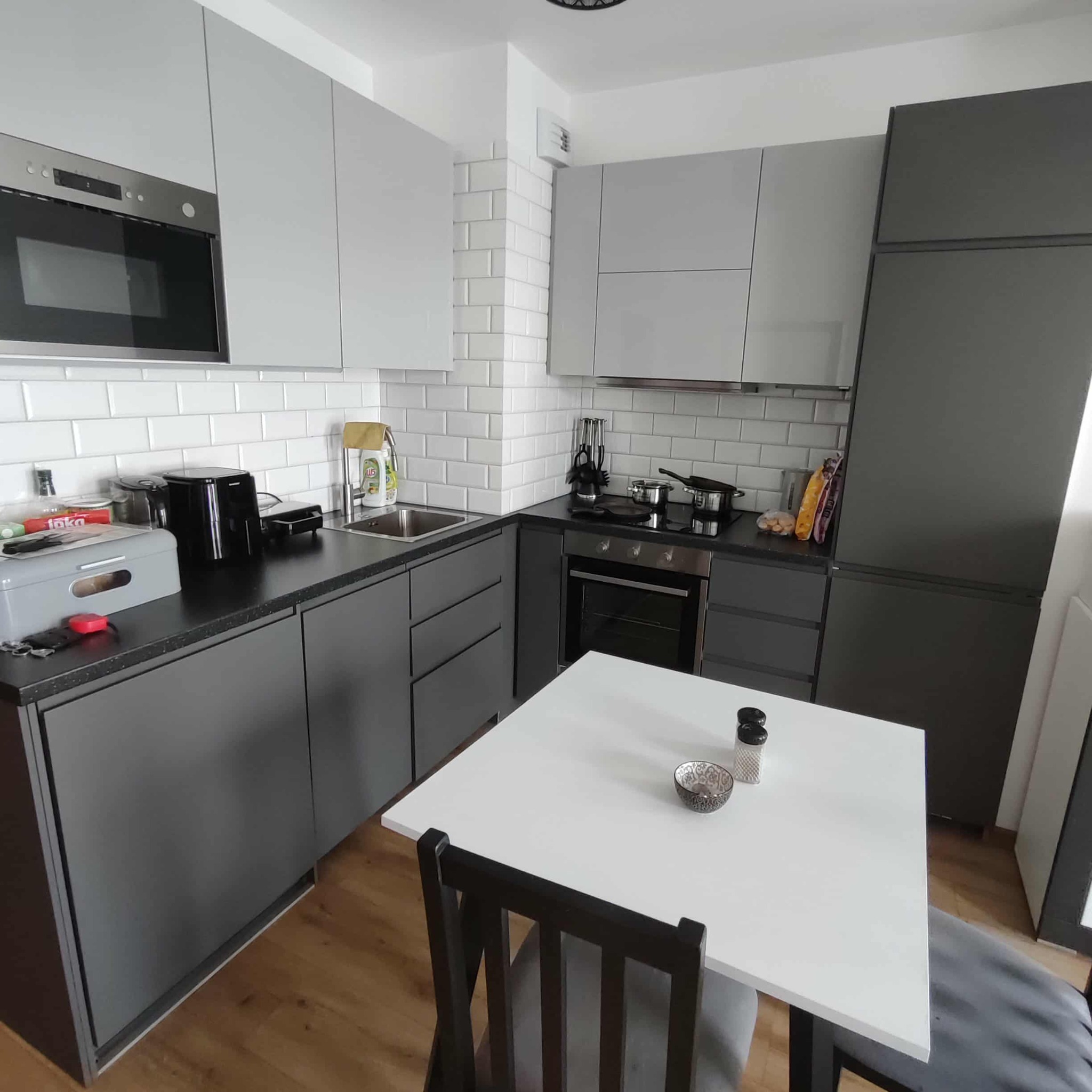 Nowoczesna, szaro-biała kuchnia z białymi płytkami na ścianie, czarnym blatem i drewnianą podłogą. Na stole miseczka i przyprawy.