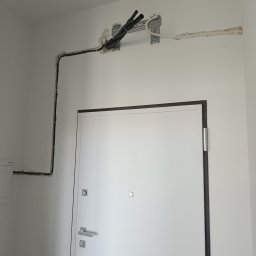 Optiklima - Białe wnętrze z widoczną instalacją klimatyzacji: rury prowadzone po ścianie, zamaskowane gipsem. Białe ściany i drzwi wejściowe. Widoczne mocowania instalacji.