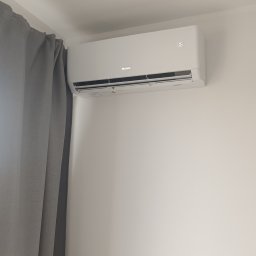 Optiklima - Biała klimatyzacja marki Gree zamontowana na ścianie w jasnym pokoju, z widocznym grzejnikiem pod spodem i zasłoną po lewej stronie. Wyświetlacz klimatyzacji pokazuje temperaturę.