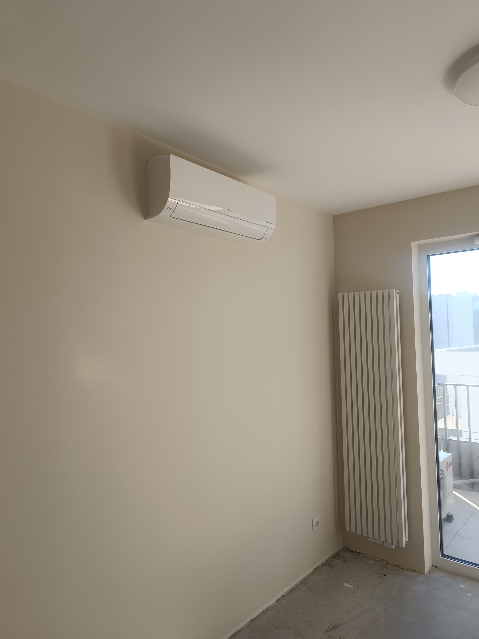 Biały klimatyzator LG zamontowany na beżowej ścianie w jasnym, minimalistycznym wnętrzu z grzejnikiem i oknem balkonowym.