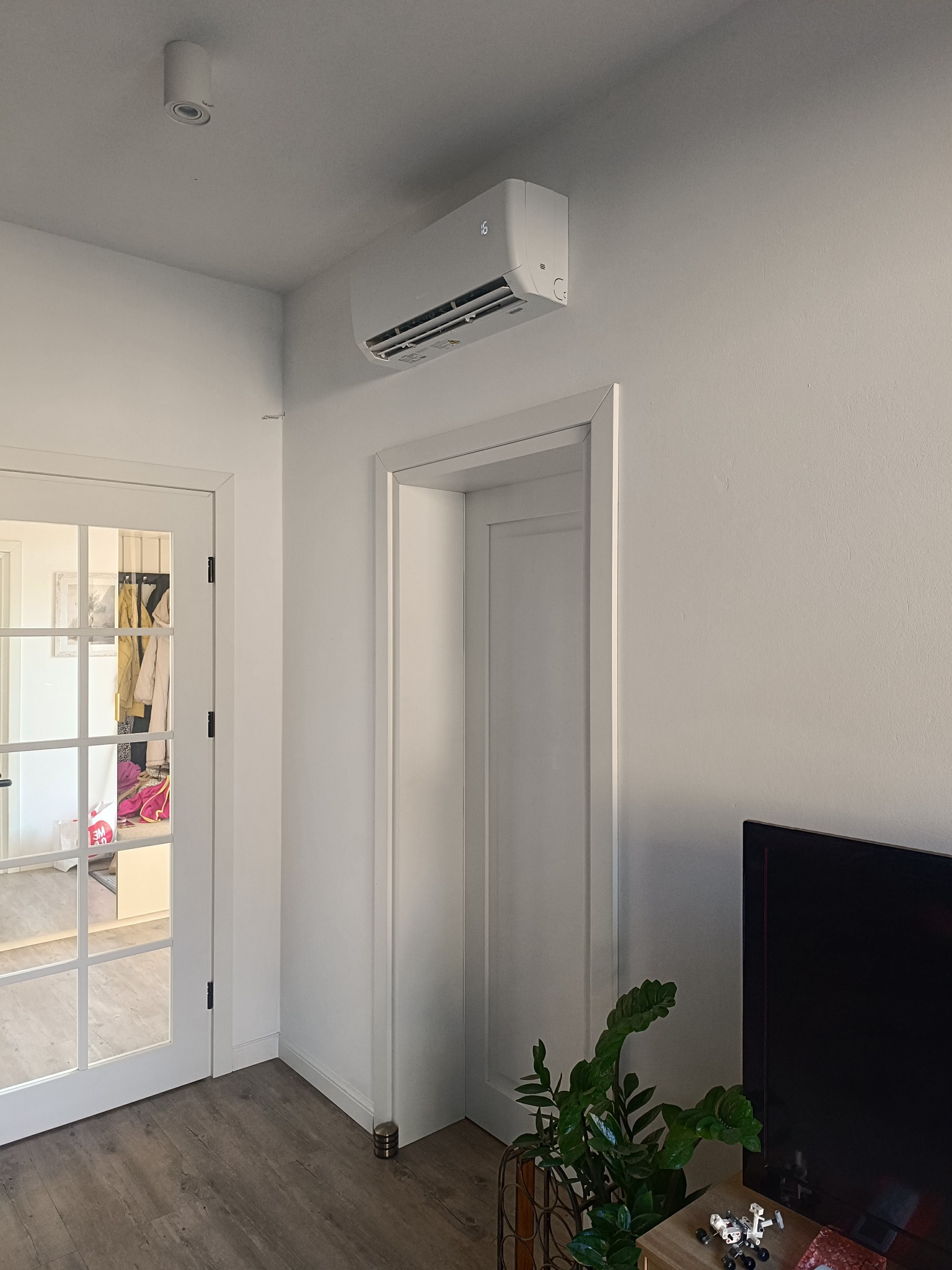 Biała klimatyzacja ścienna w nowoczesnym wnętrzu z drewnianą podłogą, widoczna nad drzwiami i obok telewizora oraz rośliny doniczkowej.