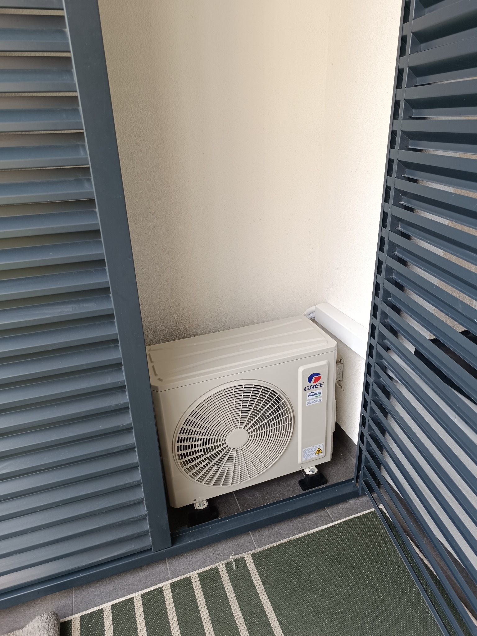 Zewnętrzna jednostka klimatyzacji Gree na balkonie, widoczna przez szare żaluzje. Kompaktowy model, zamontowany na czarnych podstawkach, z fragmentem dywanika.