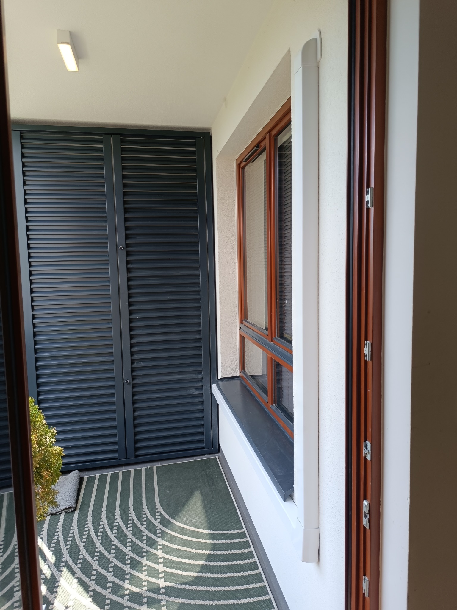 Zabudowany balkon z żaluzjami i oknem. Widoczna instalacja klimatyzacji w białej obudowie przy oknie. Na podłodze dywanik.