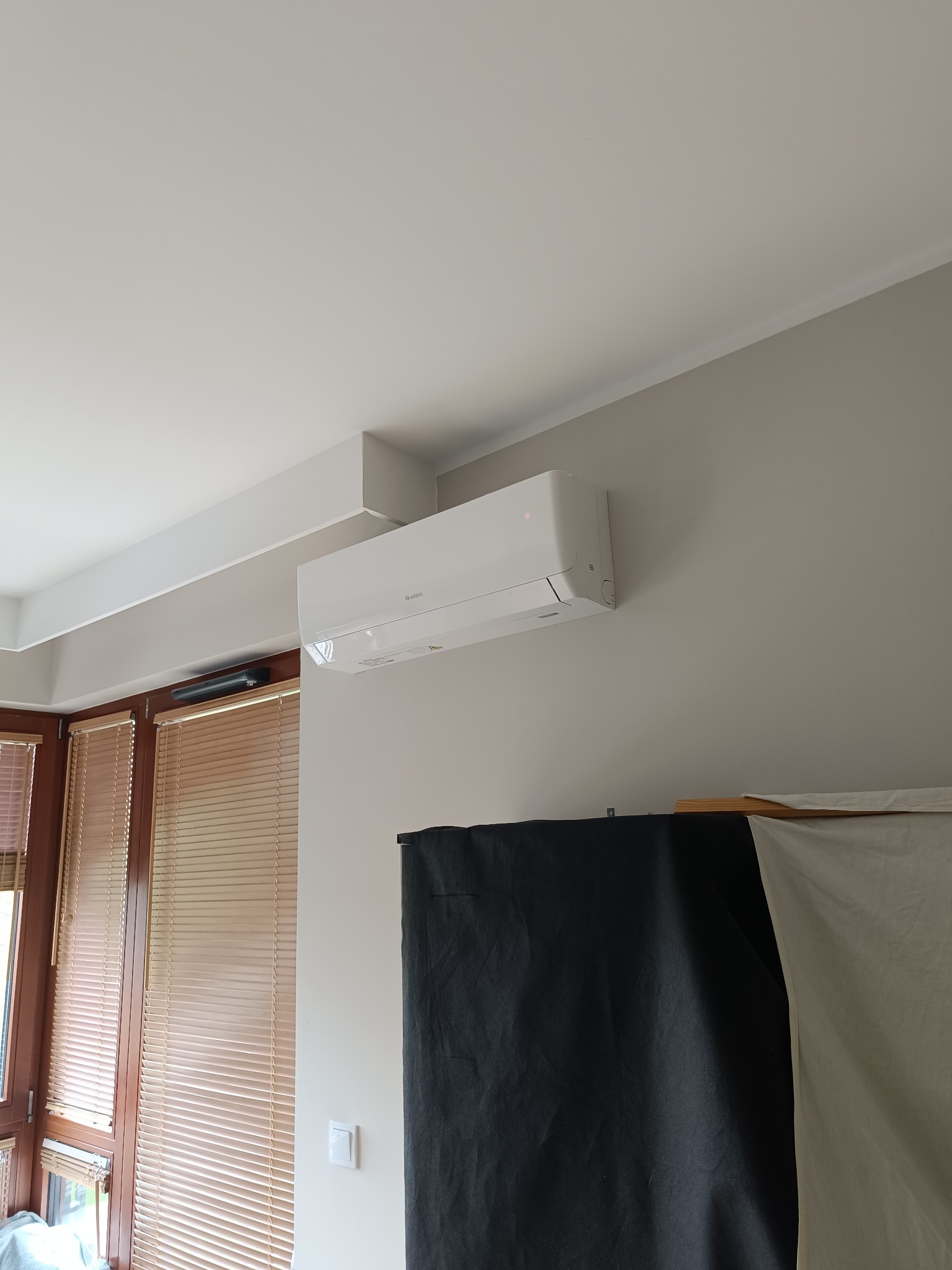 Biały klimatyzator ścienny Gree, zamontowany w jasnym pomieszczeniu z żaluzjami. Widoczny fragment czarnego materiału w tle.
