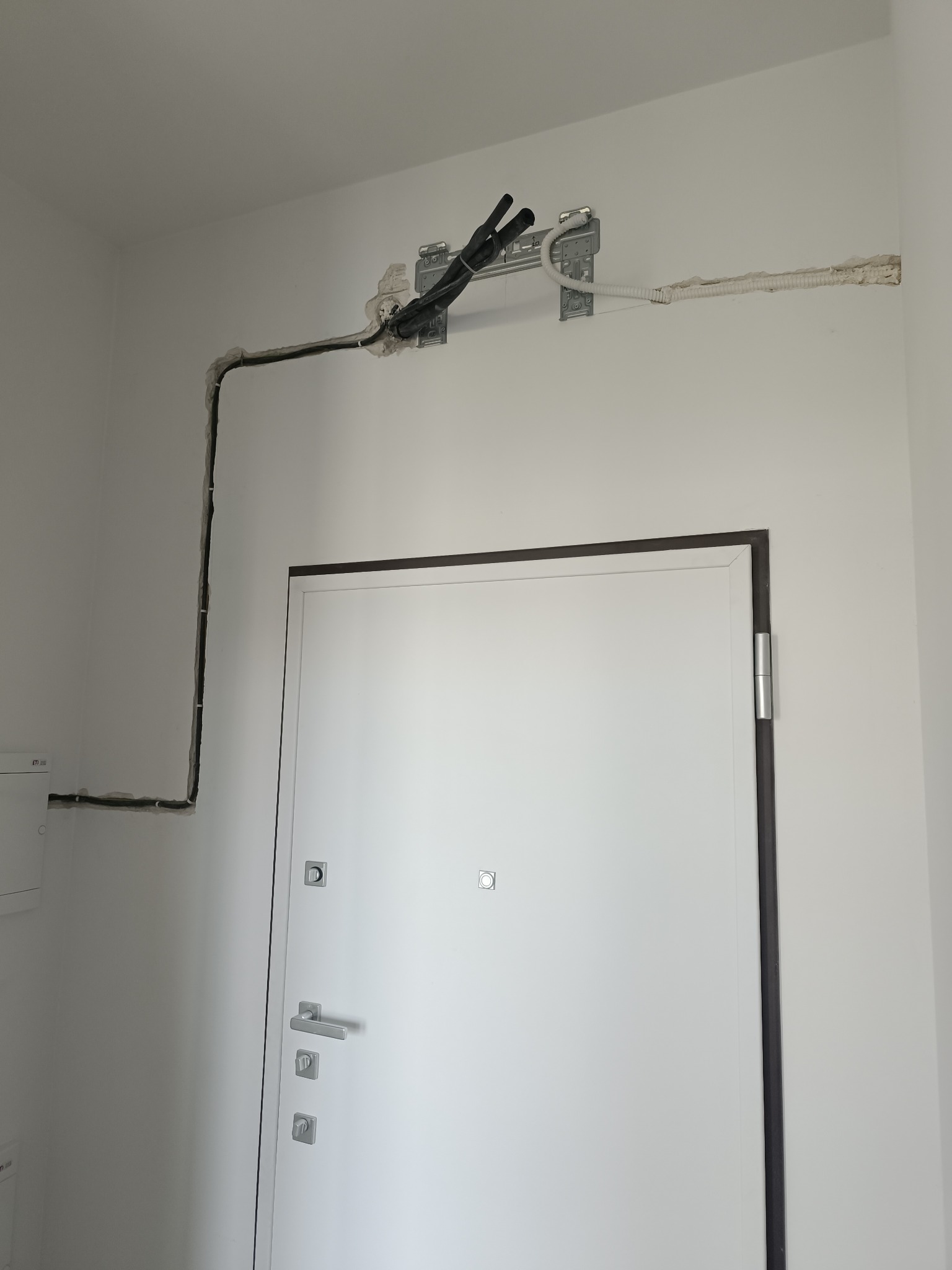 Białe wnętrze z widoczną instalacją klimatyzacji: rury prowadzone po ścianie, zamaskowane gipsem. Białe ściany i drzwi wejściowe. Widoczne mocowania instalacji.
