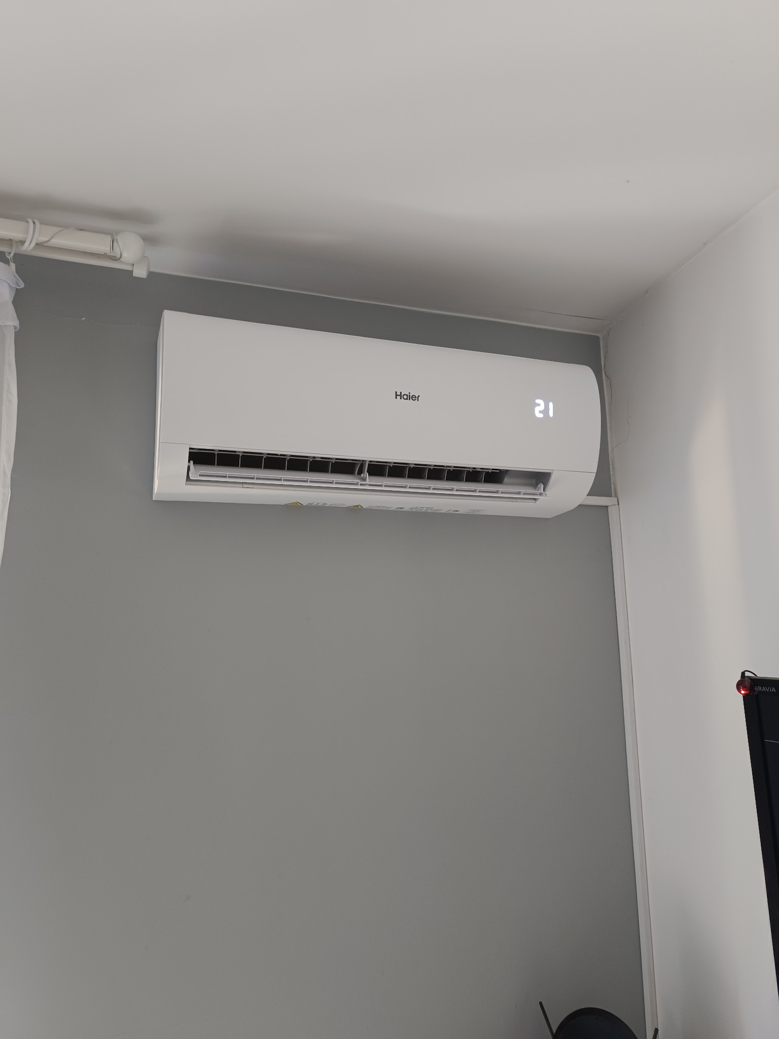 Biała klimatyzacja Haier zamontowana na szarej ścianie w pomieszczeniu. Na wyświetlaczu widoczna temperatura 21 stopni. Widoczny fragment telewizora.