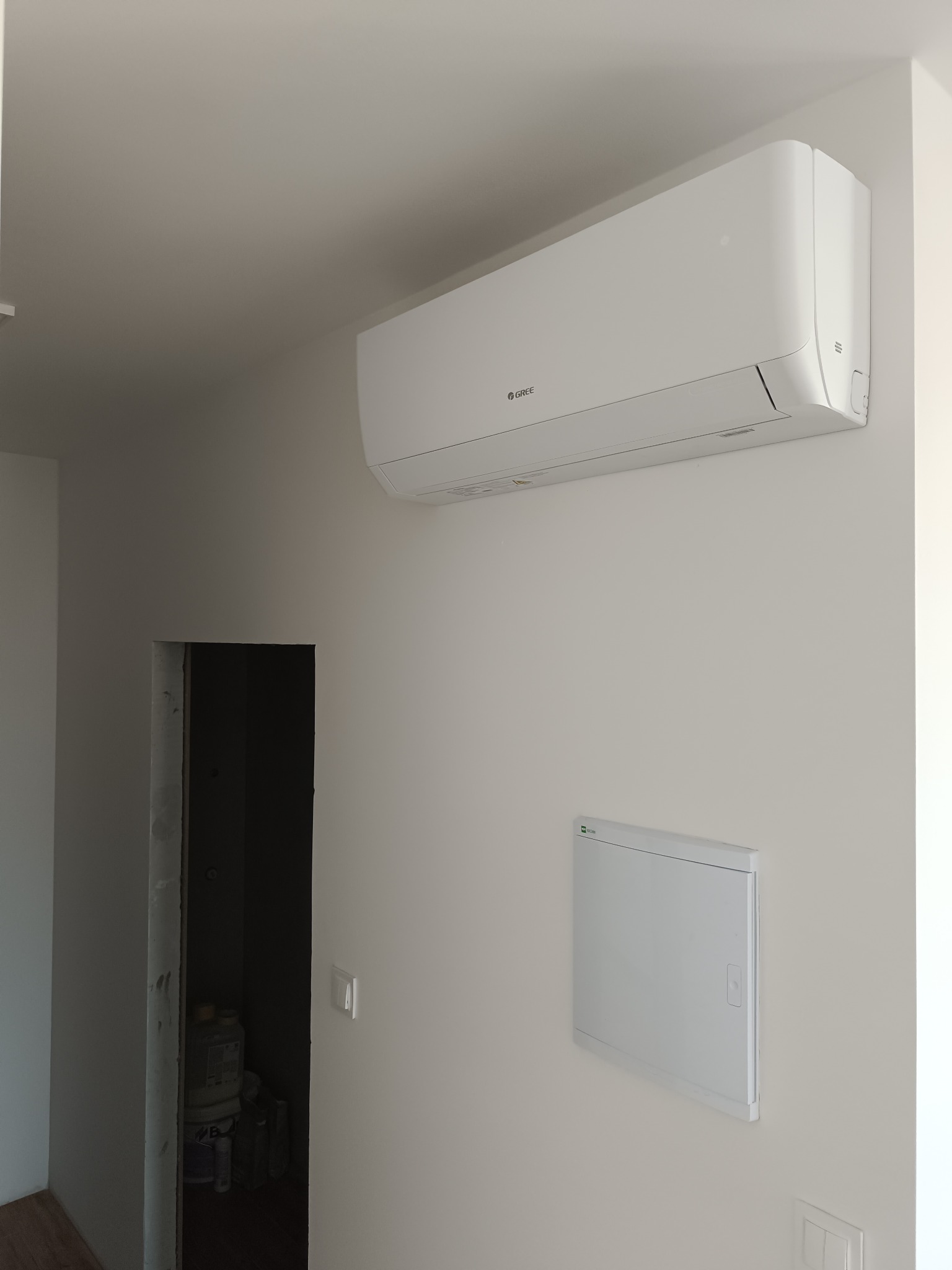 Biały klimatyzator Gree zamontowany na jasnej ścianie w nowym pomieszczeniu z widoczną instalacją elektryczną i otwartym przejściem do kolejnego pokoju.