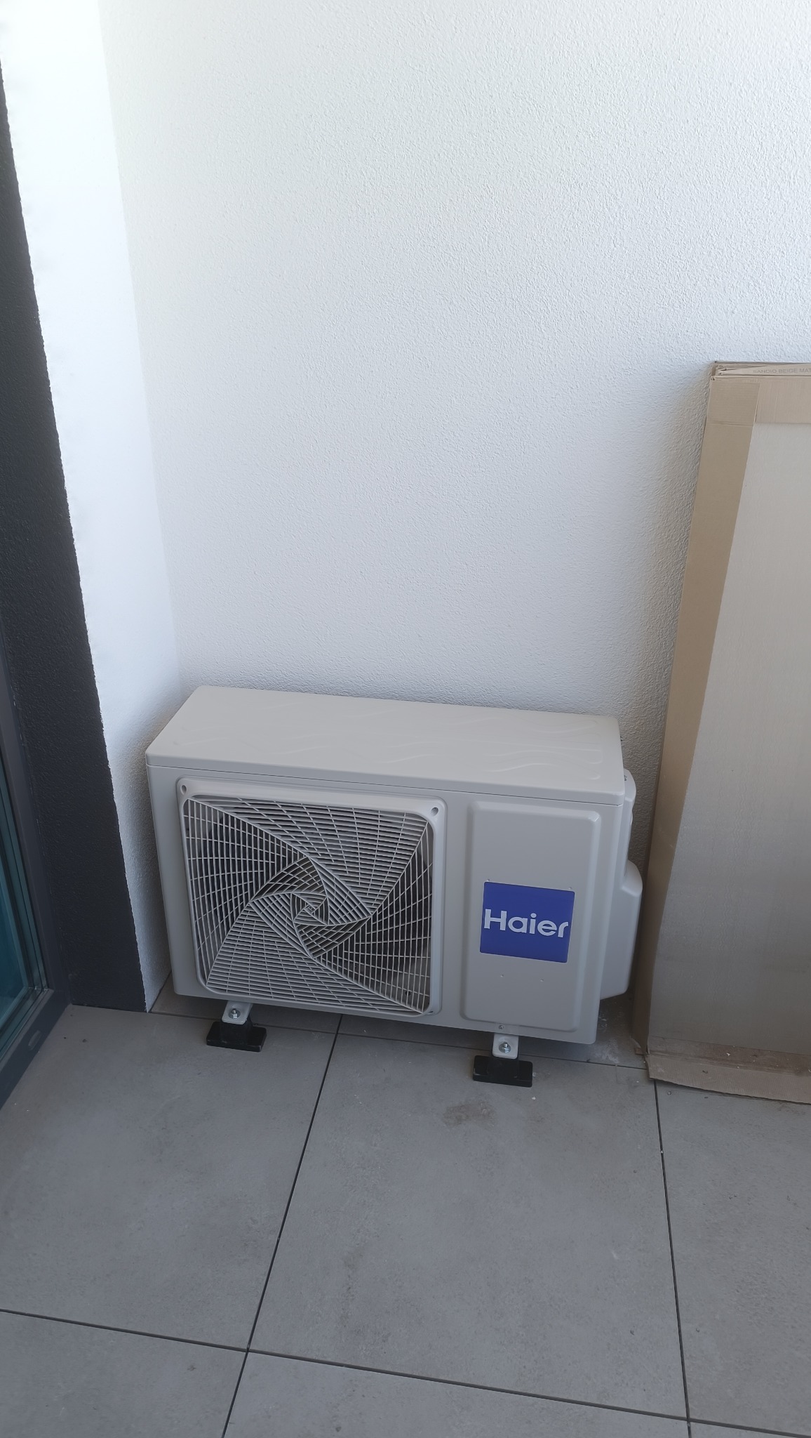 Biała, zewnętrzna jednostka klimatyzacji Haier zamontowana na szarych płytkach, przy białej ścianie. Widoczne elementy montażowe i fragment kartonu.