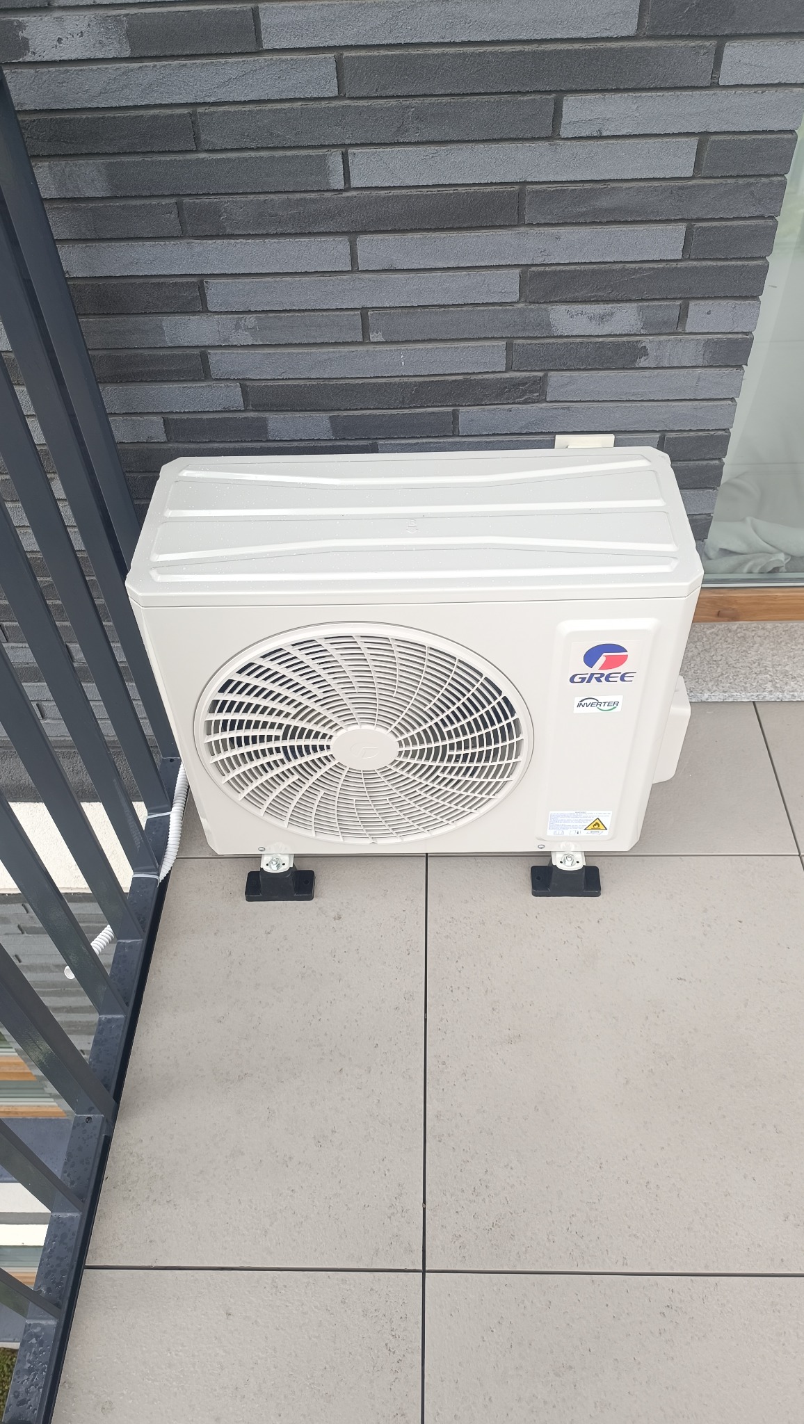 Biała jednostka zewnętrzna klimatyzacji GREE na balkonie z szarą balustradą i płytkami. Widoczna ściana z szarej cegły.