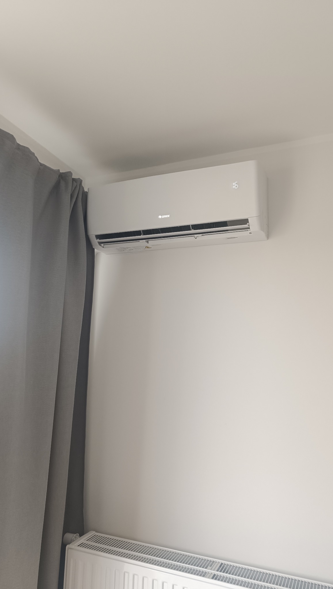 Biała klimatyzacja marki Gree zamontowana na ścianie w jasnym pokoju, z widocznym grzejnikiem pod spodem i zasłoną po lewej stronie. Wyświetlacz klimatyzacji pokazuje temperaturę.