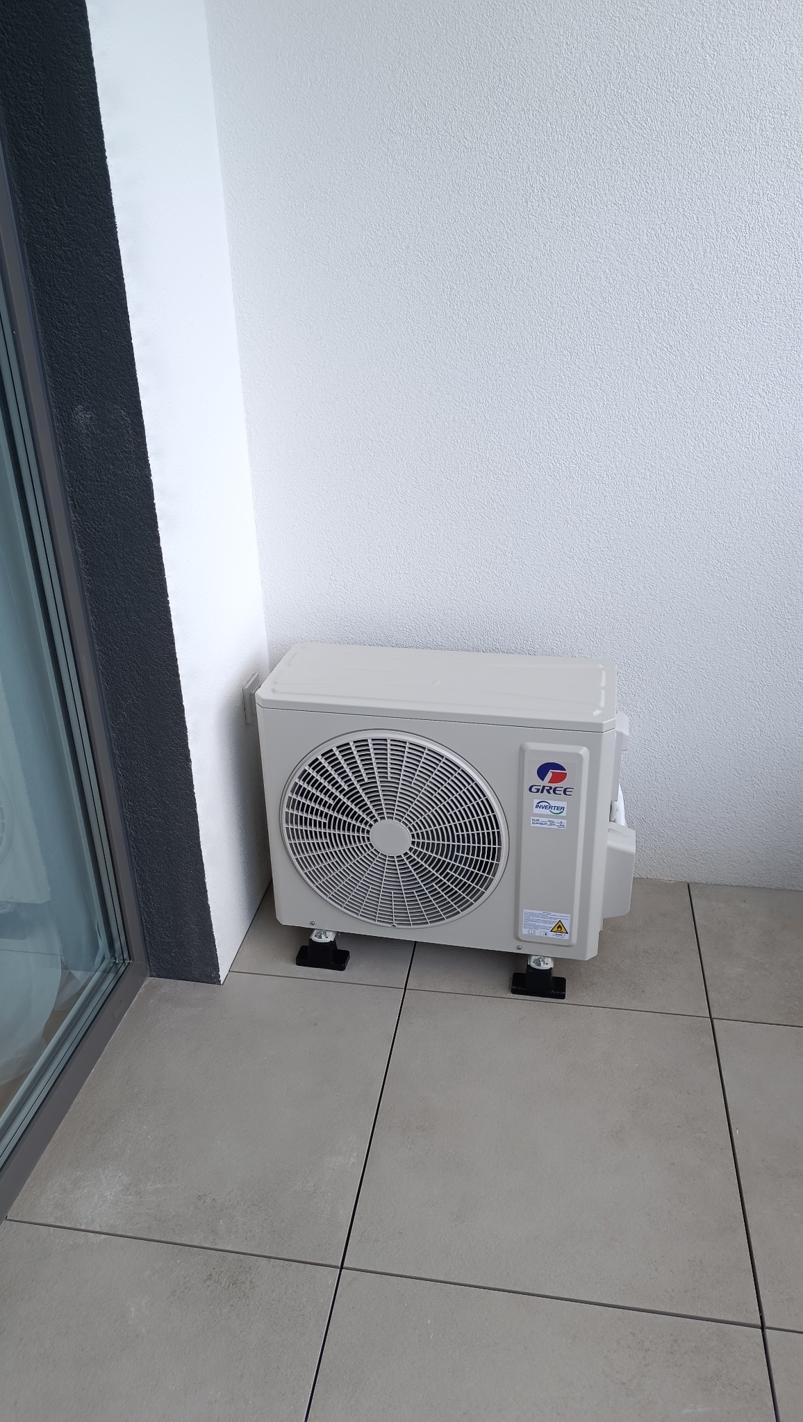 Kompaktowa jednostka zewnętrzna klimatyzacji GREE Inverter na balkonie, zamontowana na czarnych podstawkach antywibracyjnych. Minimalistyczny design, jasne ściany i szara podłoga.