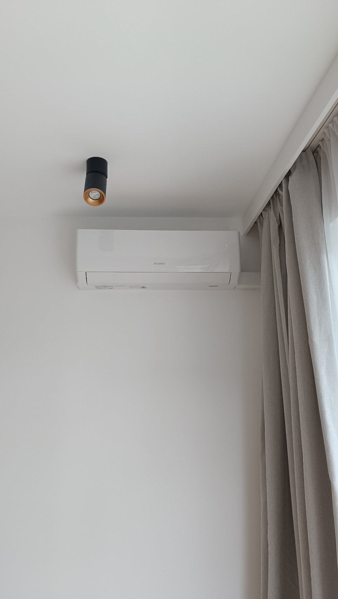 Biały klimatyzator Gree zamontowany na ścianie, obok lampa sufitowa i zasłony. Minimalistyczny design wnętrza, stonowana kolorystyka.