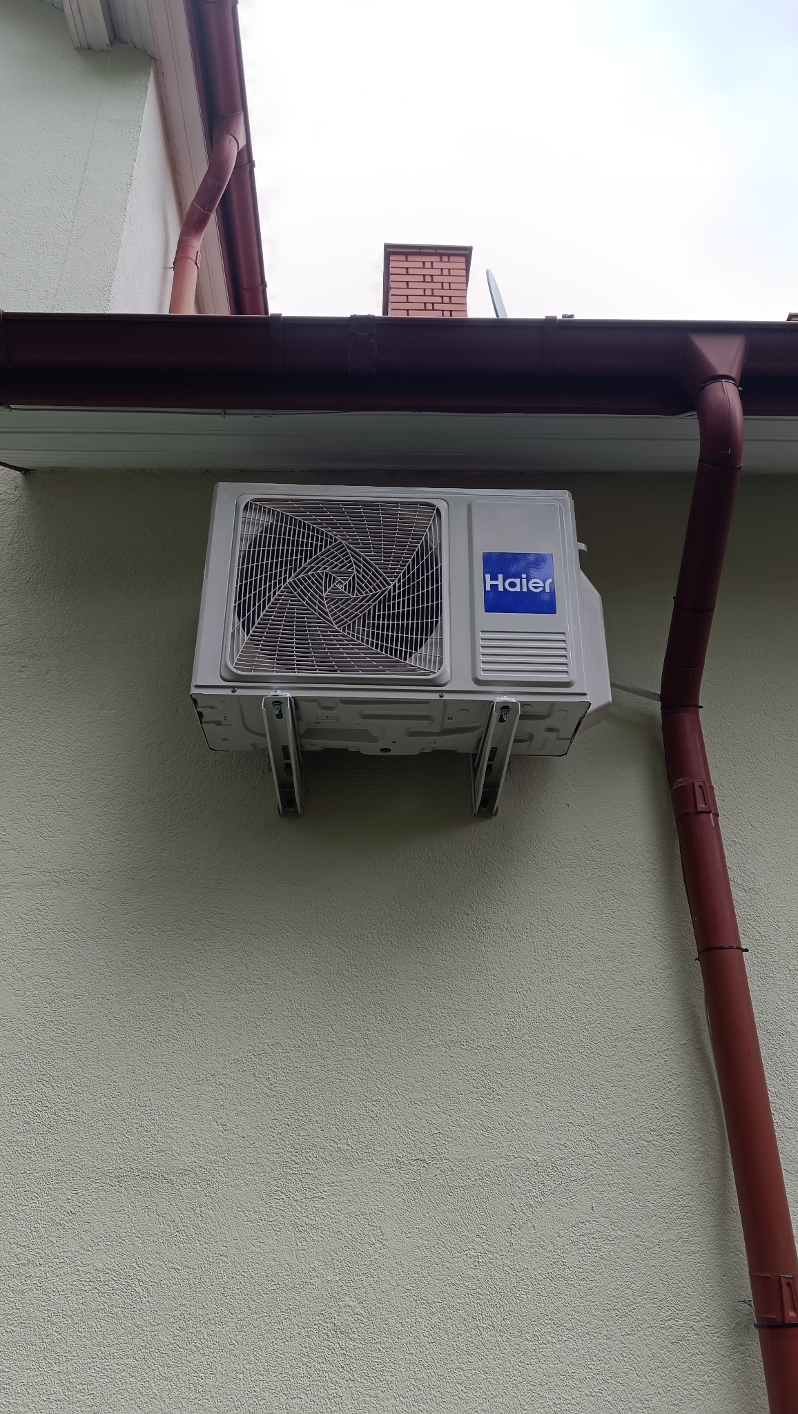 Zamontowana jednostka zewnętrzna klimatyzacji Haier na elewacji budynku z brązową rynną i kominem w tle, widok z dołu.
