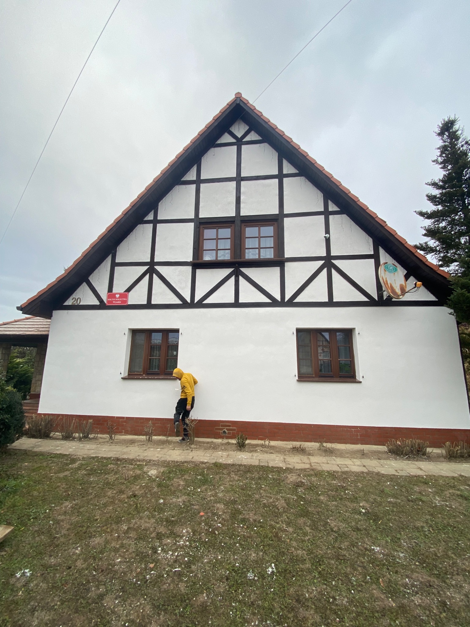 Malowanie elewacji domu w stylu szachulcowym w Rudnej. Osoba w żółtej bluzie maluje białą farbą ścianę. Widoczny numer domu i antena satelitarna.