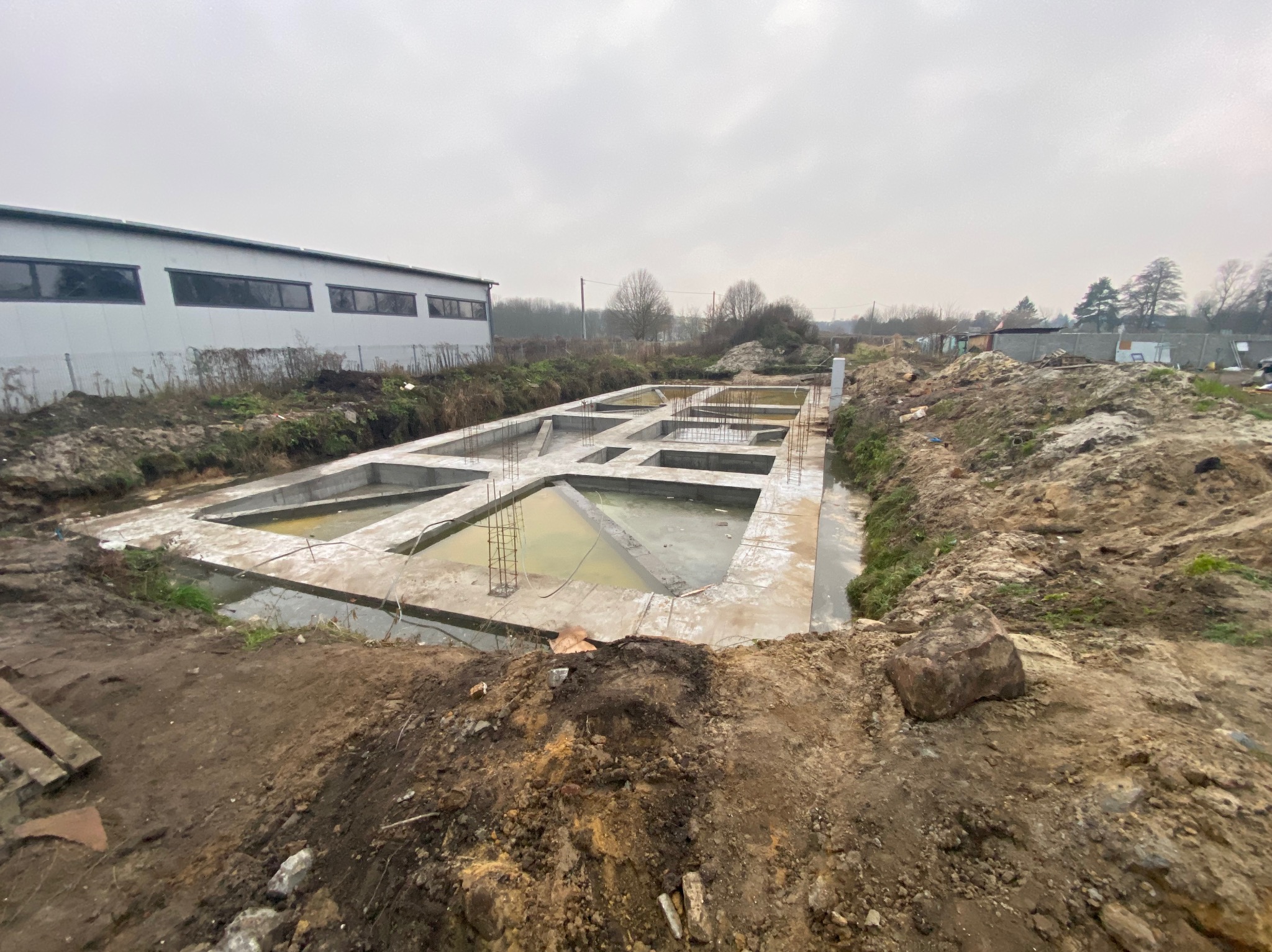 Wylane fundamenty pod budynek z widocznym zbrojeniem, częściowo wypełnione wodą. W tle budynek przemysłowy i drzewa. Pochmurne niebo.
