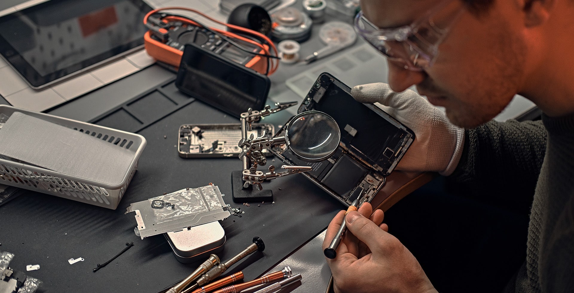 Serwisant w okularach, w rękawicy, naprawia otwarty smartfon na czarnym stole warsztatowym przy użyciu precyzyjnego śrubokręta i lupy na statywie; widoczne narzędzia, części zamienne i tablet w tle.