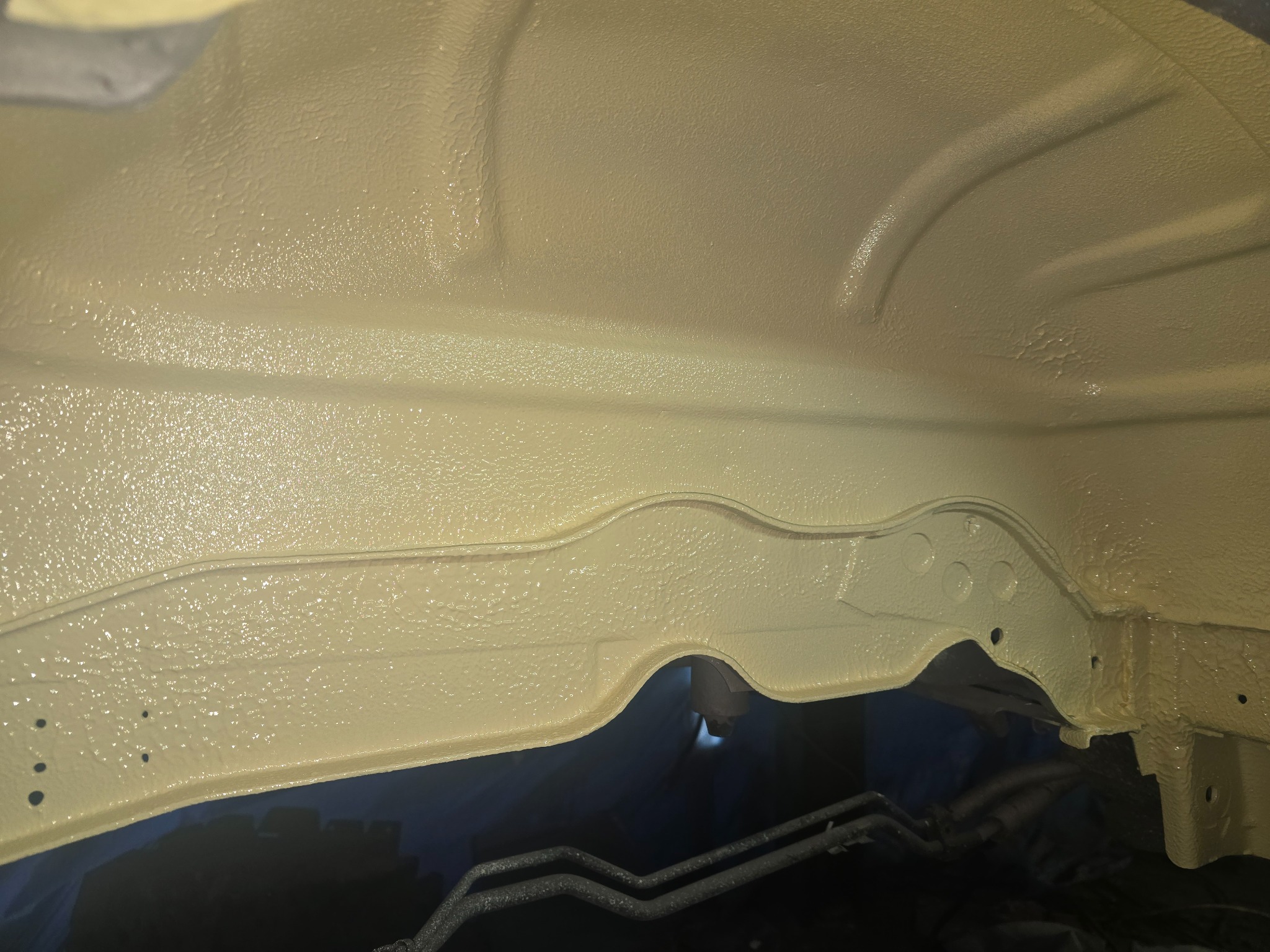 Podwozie samochodu po nałożeniu ochronnej warstwy antykorozyjnej w kolorze beżowym, widoczne przewody paliwowe i elementy konstrukcyjne.