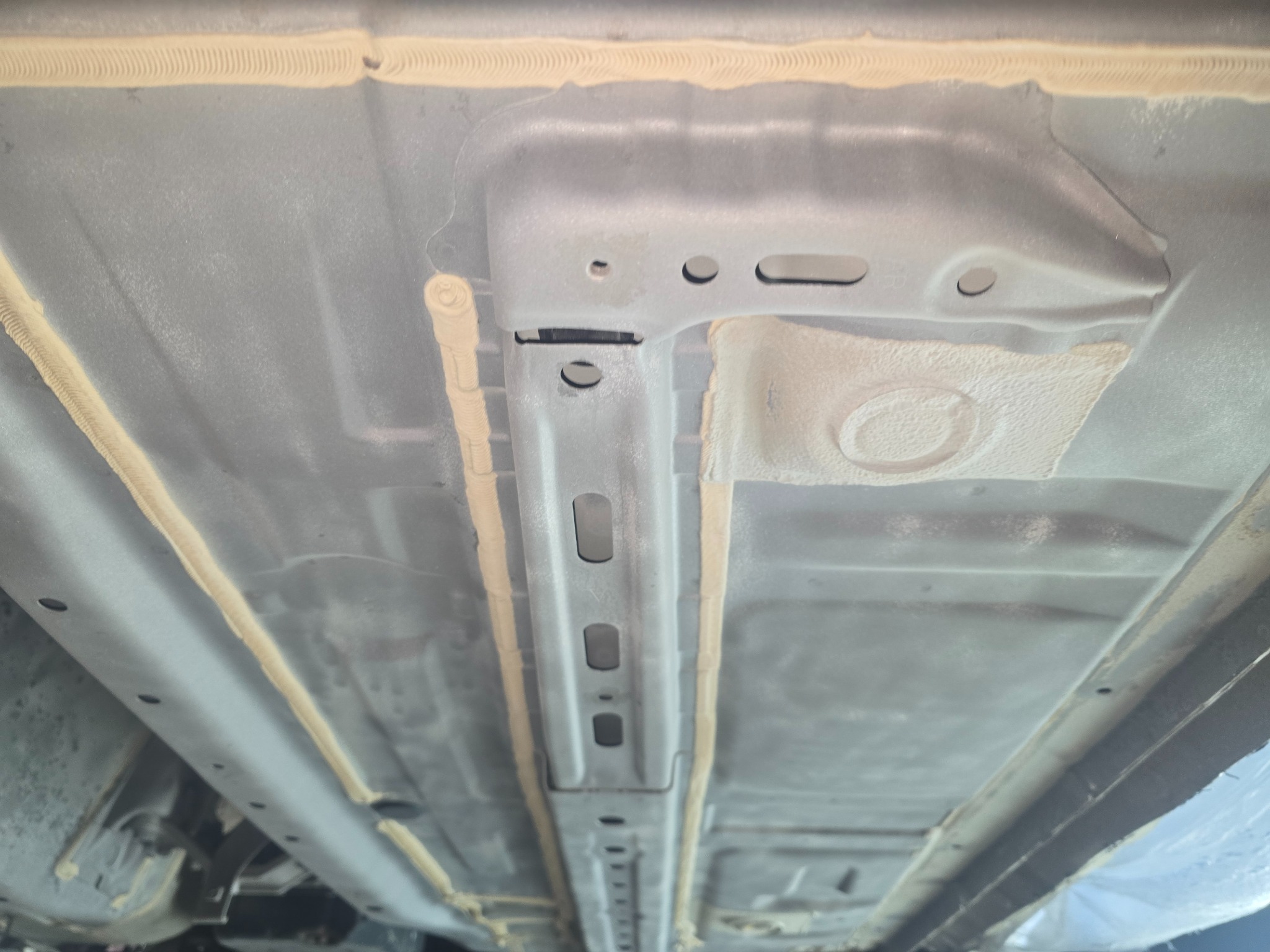 Spód samochodu po zabezpieczeniu antykorozyjnym, widoczne metalowe elementy ramy i podwozia pokryte grubą warstwą ochronną w kolorze beżowym, aplikowaną w charakterystyczny, prążkowany sposób.