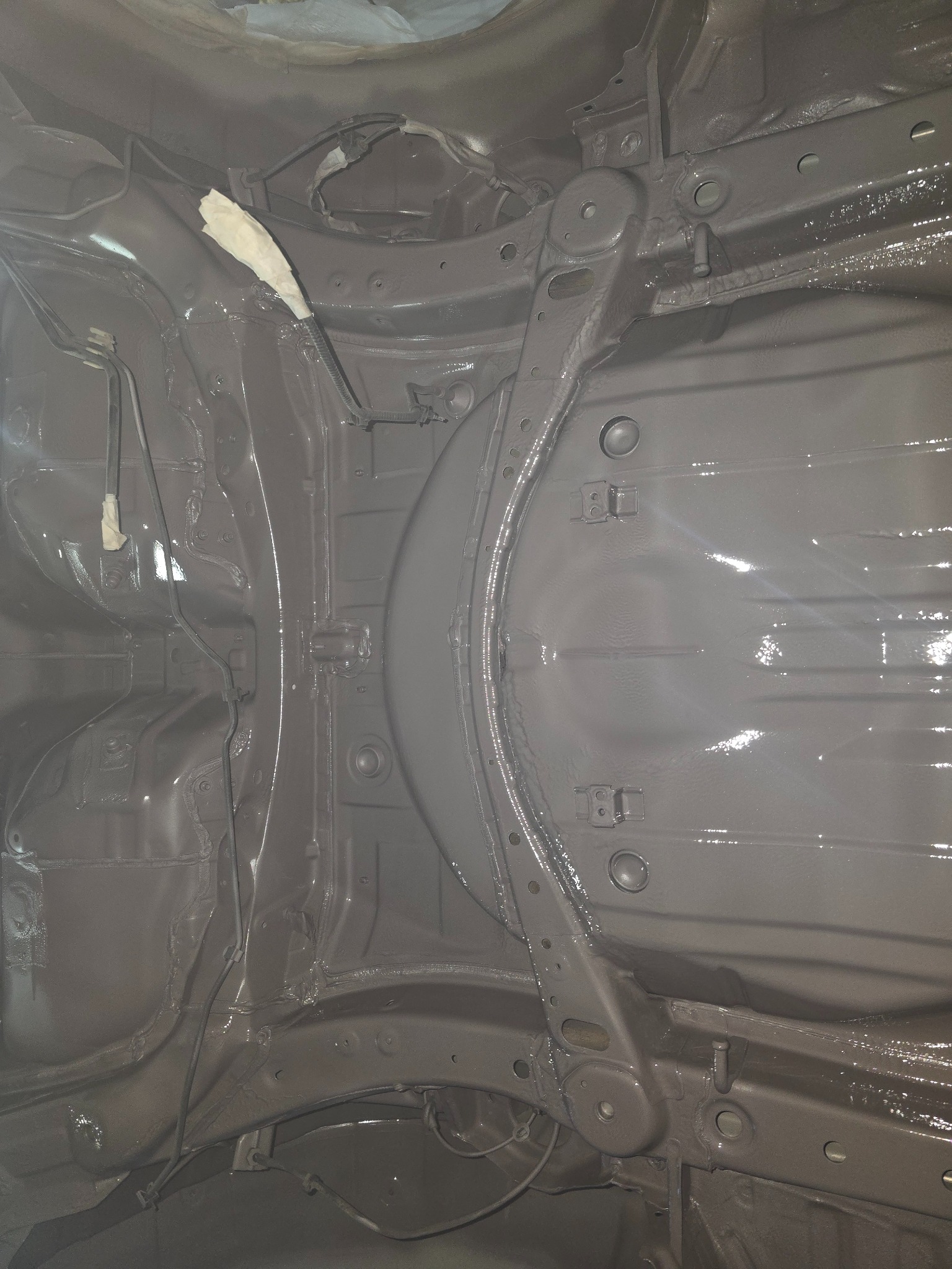 Spód samochodu po nałożeniu szarej, błyszczącej powłoki antykorozyjnej, z widocznymi elementami zawieszenia i przewodami zabezpieczonymi taśmą malarską.