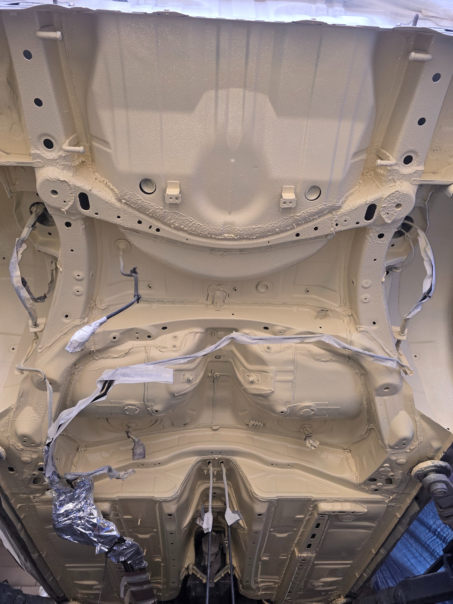 Podwozie samochodu po zabiegu antykorozyjnym, widoczne elementy zabezpieczone folią i taśmą maskującą, świeżo nałożona warstwa ochronna w kolorze beżowym.