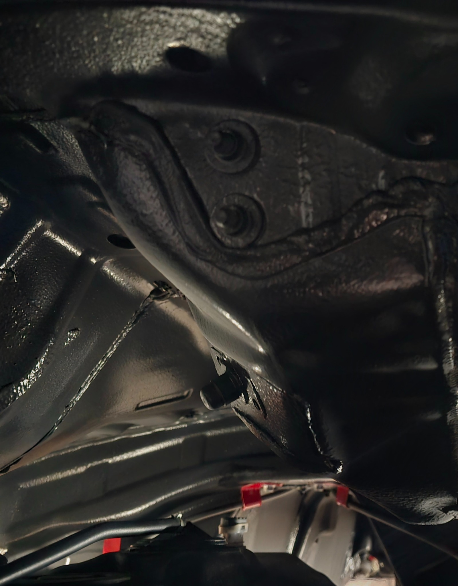 Podwozie samochodu po zabezpieczeniu antykorozyjnym, widoczne elementy metalowe pokryte czarną, błyszczącą powłoką, śruby i fragmenty zawieszenia w dolnej części kadru.