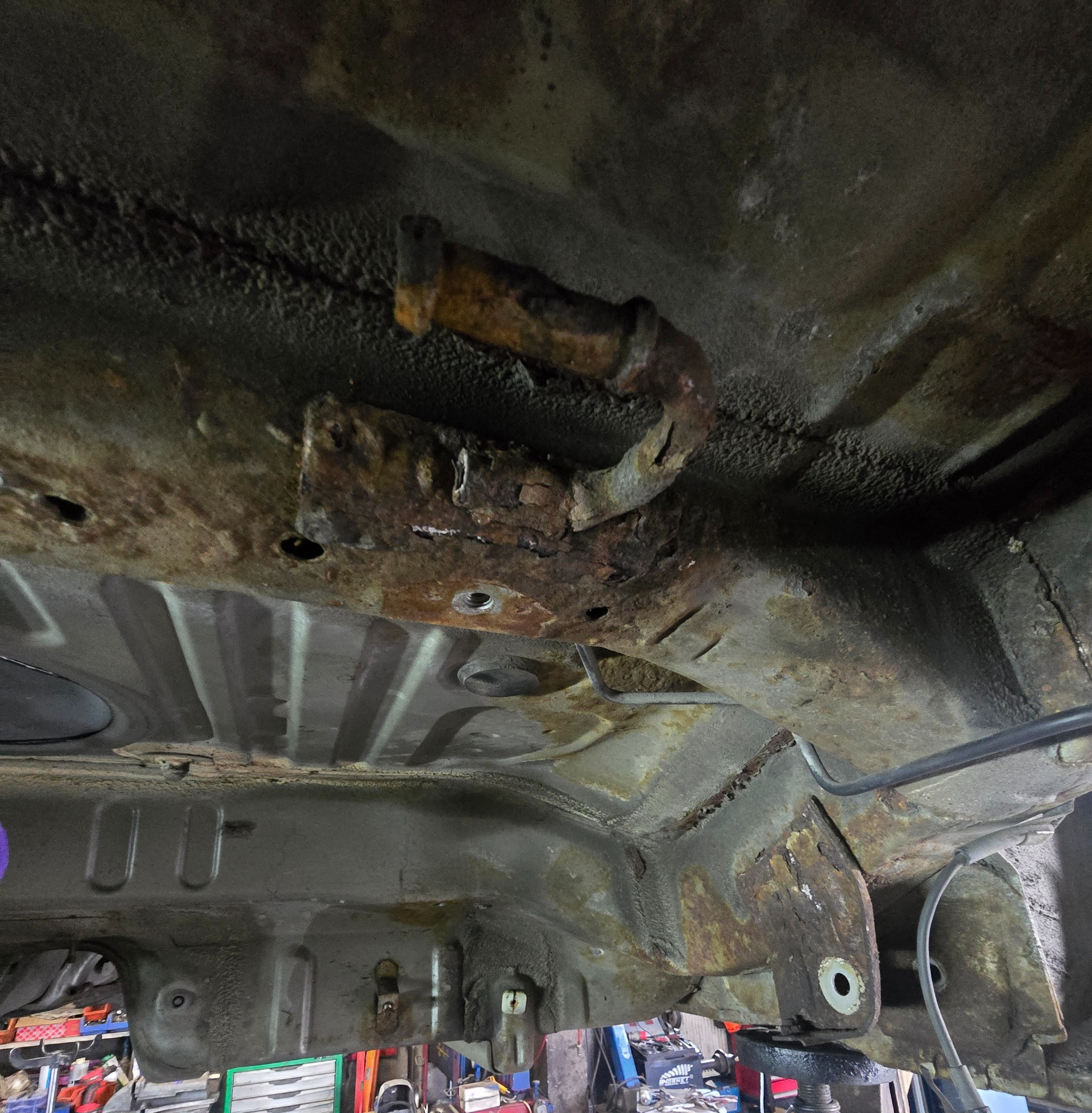 Zardzewiałe podwozie samochodu z widocznymi śladami korozji i ubytkami, sfotografowane od dołu w warsztacie. Widoczne elementy konstrukcyjne i przewody.