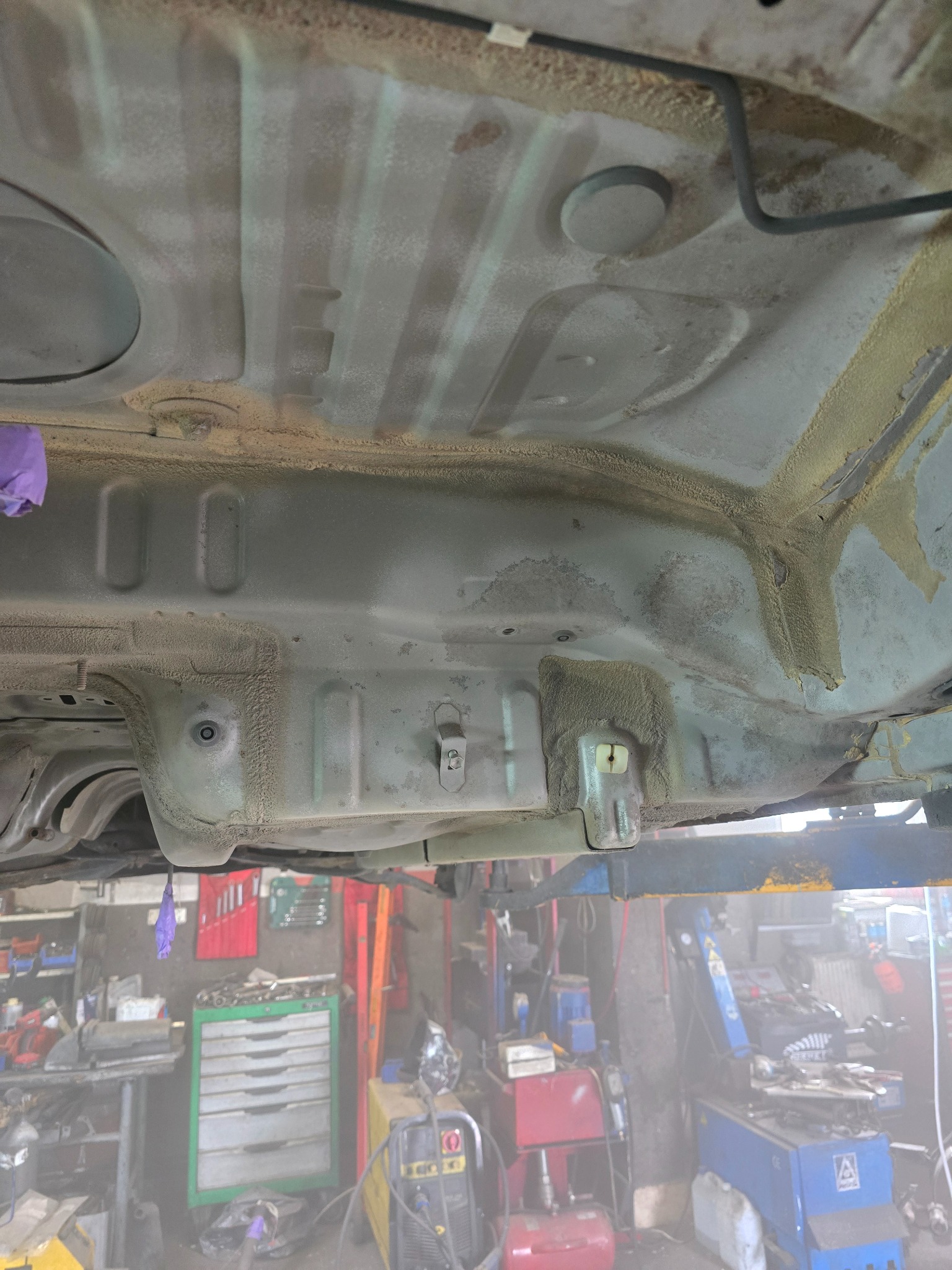 Spód samochodu na podnośniku w warsztacie, widoczne ślady korozji i zabezpieczenia antykorozyjne. W tle narzędzia i wyposażenie warsztatowe.