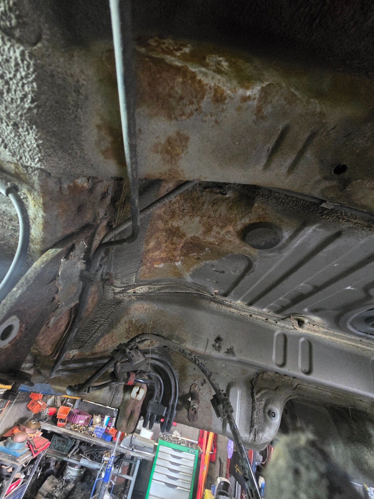 Spód skorodowanego pojazdu, widoczne przewody i fragment wyposażenia warsztatu w tle. Ukazana korozja i stan podwozia przed konserwacją.