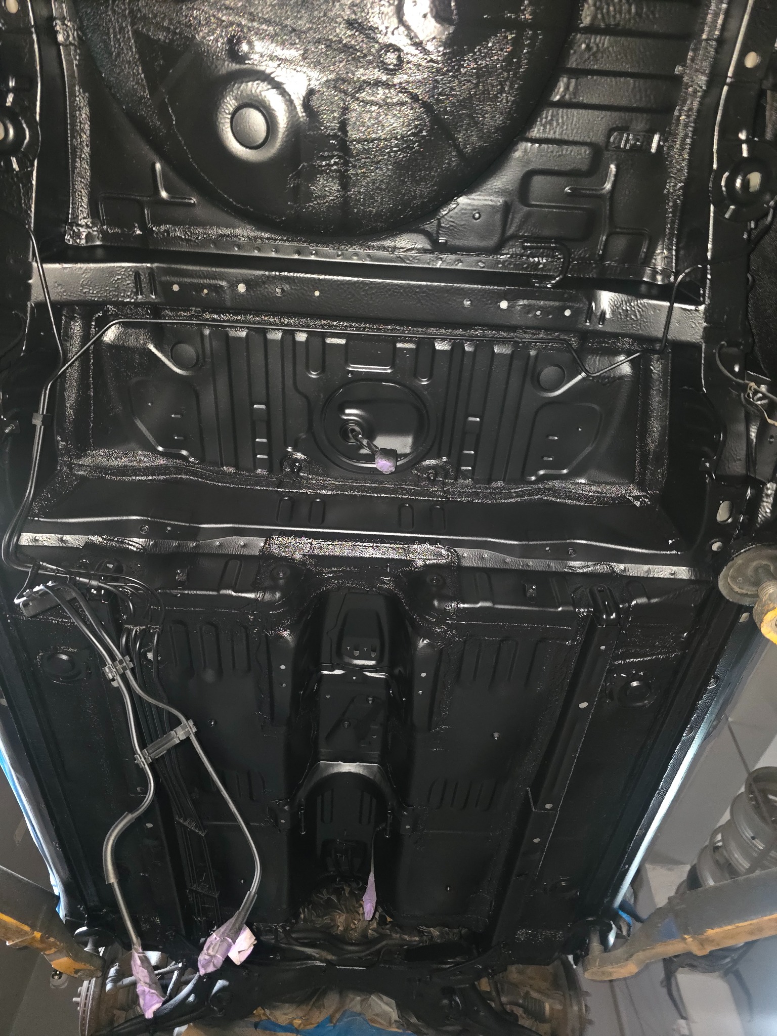 Podwozie auta po konserwacji antykorozyjnej, widoczne z dołu na podnośniku. Całość pokryta grubą, czarną warstwą ochronną. Elementy zawieszenia zabezpieczone.