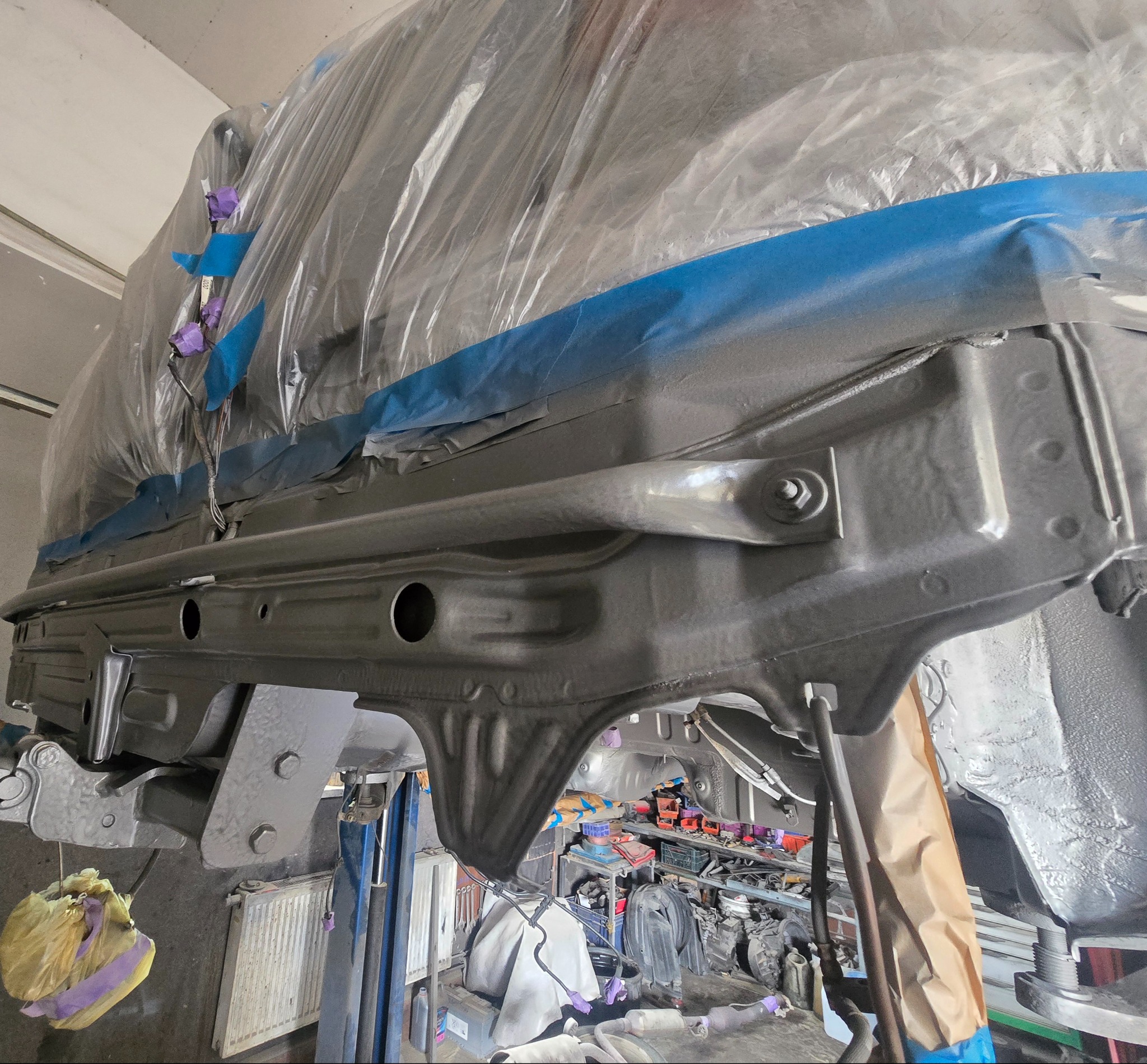 Podwozie auta po konserwacji antykorozyjnej w Łękawicy, zabezpieczone folią i taśmą. Widoczne elementy metalowe pokryte szarą farbą. Auto na podnośniku.