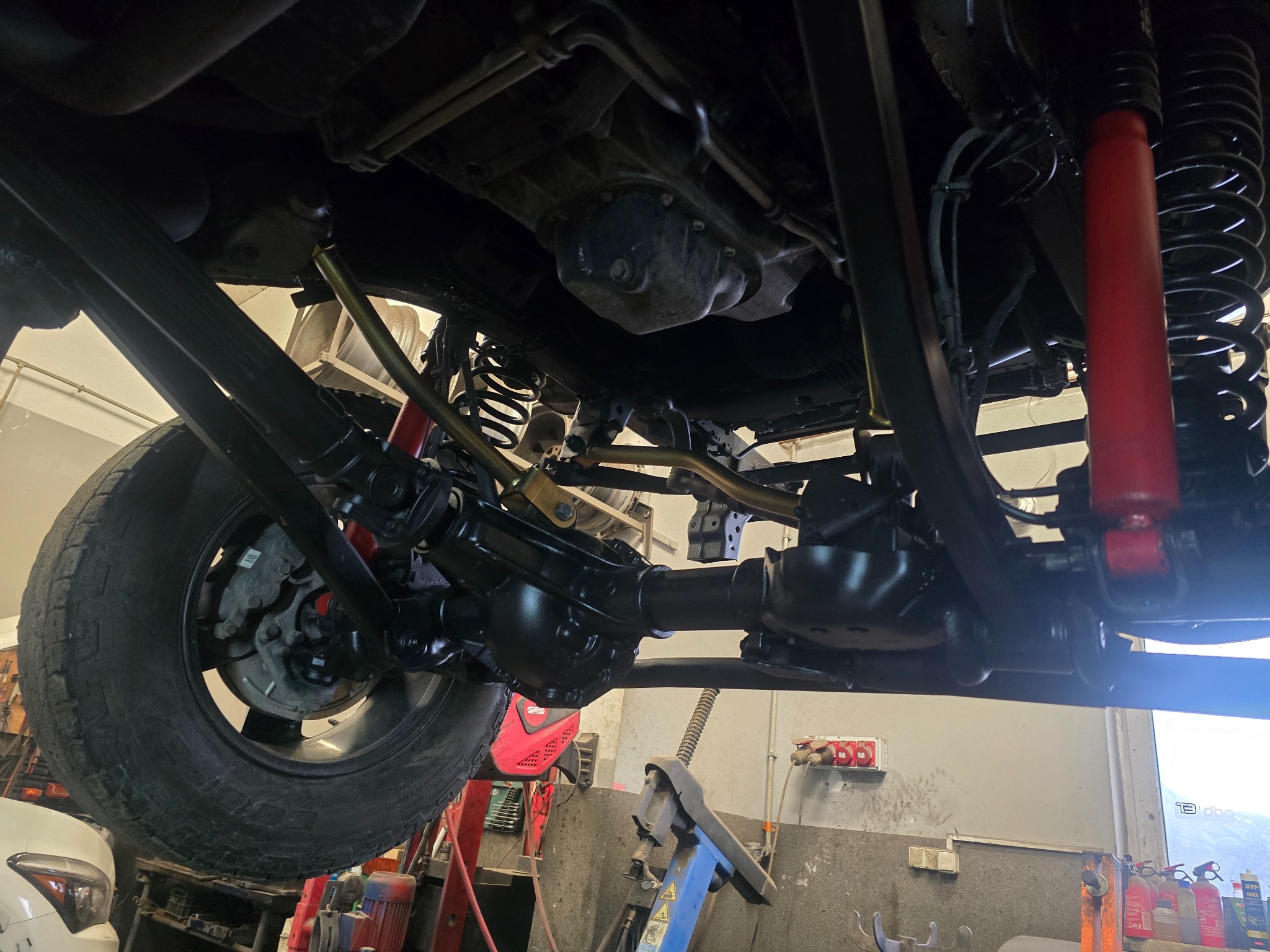 Podwozie samochodu terenowego na podnośniku, widoczne elementy zawieszenia, sprężyny i amortyzatory w kolorze czerwonym. Detale po konserwacji, czyste i zabezpieczone.