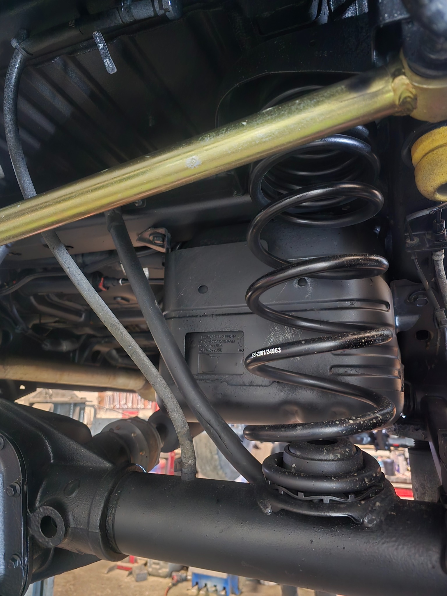 Zawieszenie pojazdu od spodu: czarna sprężyna, złoty stabilizator, elementy układu jezdnego. Widoczne oznaczenia i numery seryjne na elementach. Detal mechaniki samochodowej.